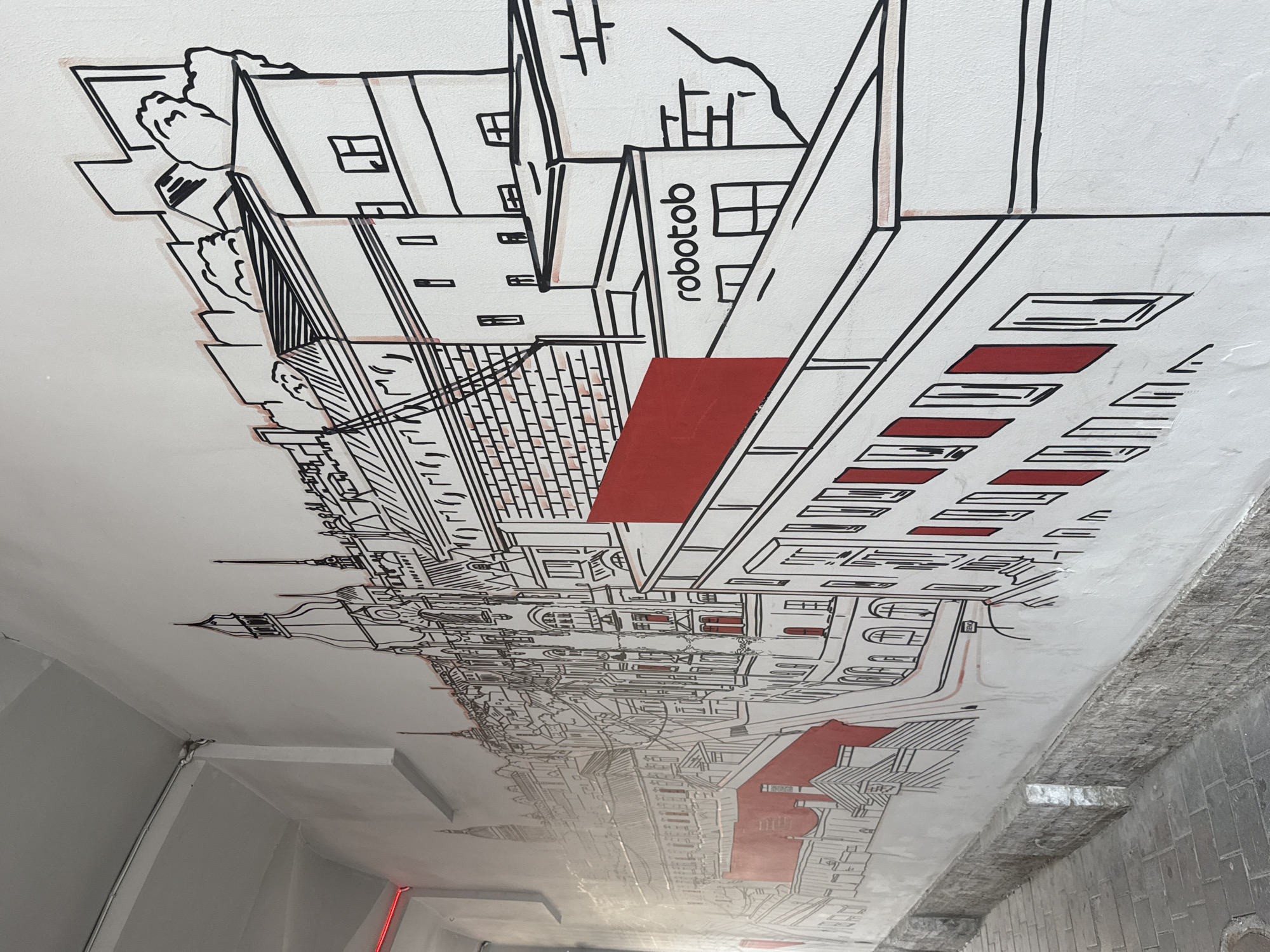 The image depicts a mural on a white wall in an outdoor urban setting. The mural is a black and white line drawing of a cityscape, featuring numerous buildings with architectural details such as windows, roofs, and spires. Some elements of the mural are highlighted with red accents, including window frames and a large rectangular block. The word "robotob" is visible on one of the depicted buildings. The mural is located on the side of a narrow walkway, with a cobblestone path visible in the foreground. The overall impression is one of urban art integrated into the city's architecture. Based on the architectural style of the mural, it is likely located in the city of Saint Petersburg, Russia.