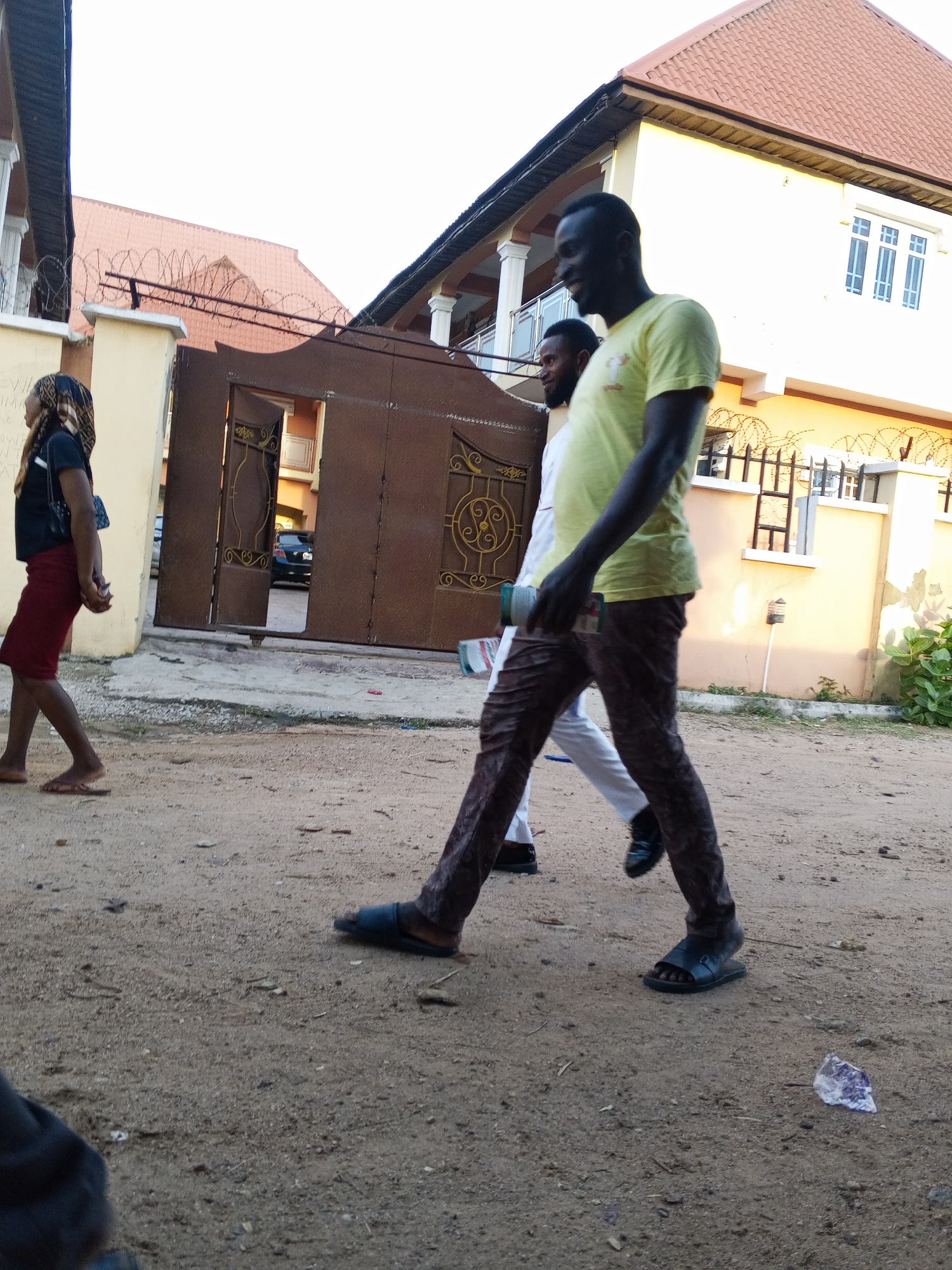 This image captures a street scene in Adonkolo, Nigeria, featuring three individuals walking past residential buildings. The time of day appears to be bright, possibly late afternoon, given the clear lighting.
In the foreground and center, a dark-skinned man wearing a yellow t-shirt and dark, patterned trousers walks towards the right, looking back over his left shoulder with a slight smile. He is wearing dark sandals and holds a green cylindrical object in his left hand. Behind him, another dark-skinned man in a white shirt and white pants walks in the same direction, wearing dark dress shoes. On the far left, a woman, also dark-skinned, walks towards the left. She is dressed in a dark top, a reddish-brown skirt, and a patterned headscarf, carrying a small dark shoulder bag, and wearing flip-flops.
The setting is an unpaved, sandy road. To the left, a light-colored pillar and wall stand beside a large, dark brown metal gate with ornate gold-colored scrollwork, which is partially open, revealing a parked black car within the compound. Along the top of the wall and surrounding fences, strands of barbed wire are visible, common for security in residential areas. The background features several houses with light-colored walls, prominent white pillars, and reddish-brown tiled roofs. One house has distinct white-framed windows. There are some faint, illegible markings or graffiti on the light-colored wall next to the woman. A small piece of white and purple plastic debris lies on the sandy ground in the bottom right.