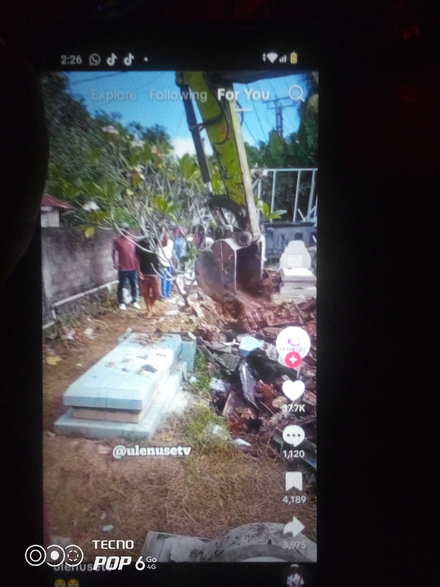 The image displays a mobile phone screen showing a TikTok video. The video appears to be filmed in an outdoor, somewhat overgrown area with what seems to be a graveyard in the foreground. There are several gravestones, one of which is partially covered by dirt. An excavator is actively digging into the ground, displacing a large amount of soil. In the background, there are a few individuals standing near a wall and some trees. The screen also shows typical TikTok interface elements such as icons for likes, comments, shares, and user engagement statistics. The time stamp at the top left indicates 2:26, and icons for WhatsApp, TikTok, and battery status are visible. The username "@ulensetv" is displayed on the screen, along with the "TECNO POP 6 4G" branding at the bottom. The location of Howayi, Nigeria, is not ascertainable from the visual information provided in the image.