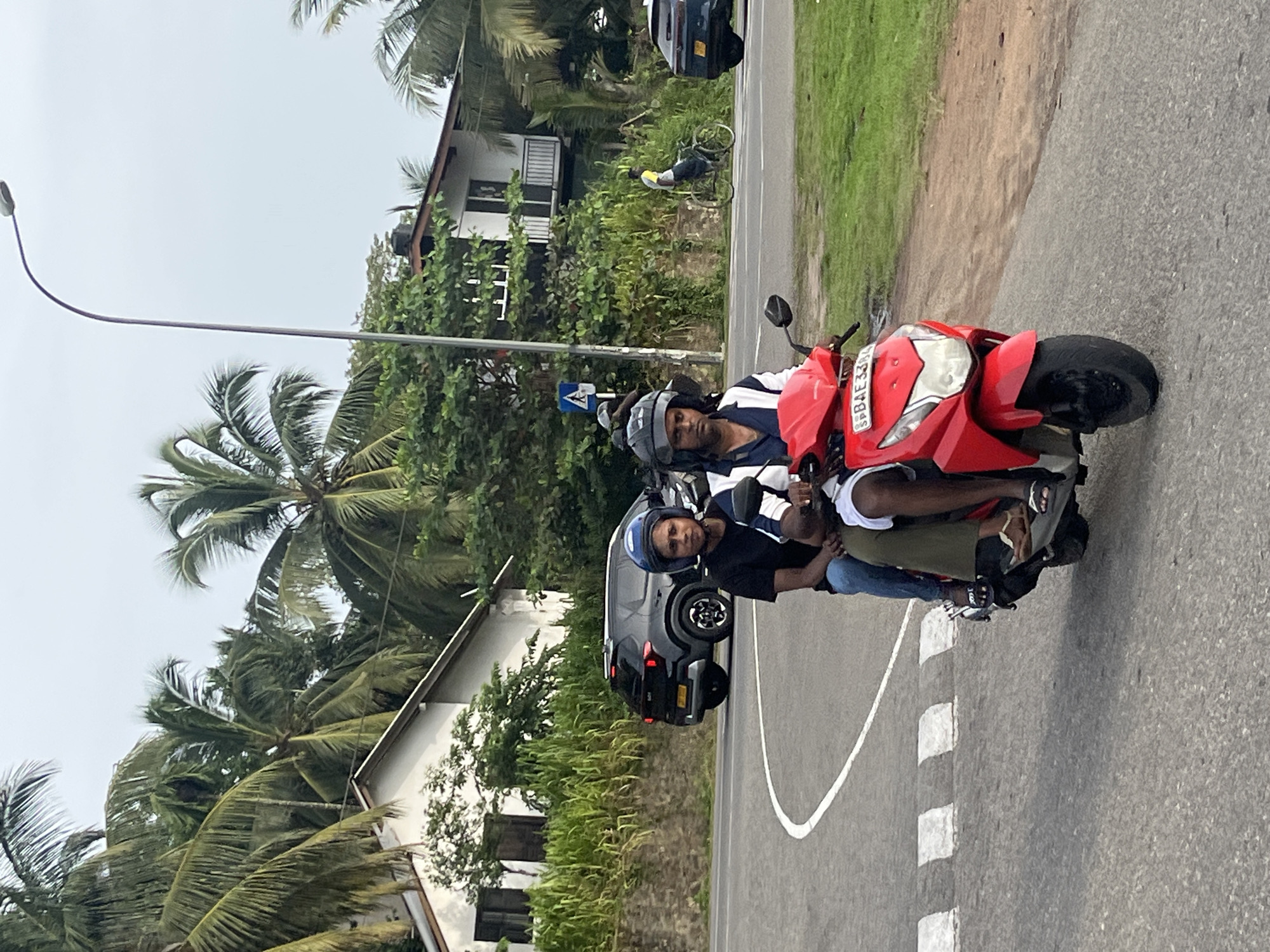 In Walahanduwa, Sri Lanka, two individuals are riding a red scooter on a paved road with white lane markings. The person in the front, wearing a black t-shirt and blue jeans, has a blue helmet. The person in the back, wearing a white and blue striped shirt, has a dark helmet. Both individuals are looking forward. A black SUV is parked on the side of the road behind them. In the background, there are houses with white walls and tiled roofs, surrounded by lush green vegetation and palm trees. A lamppost stands tall to the right of the frame, and a cyclist is visible further down the road. The sky is overcast.