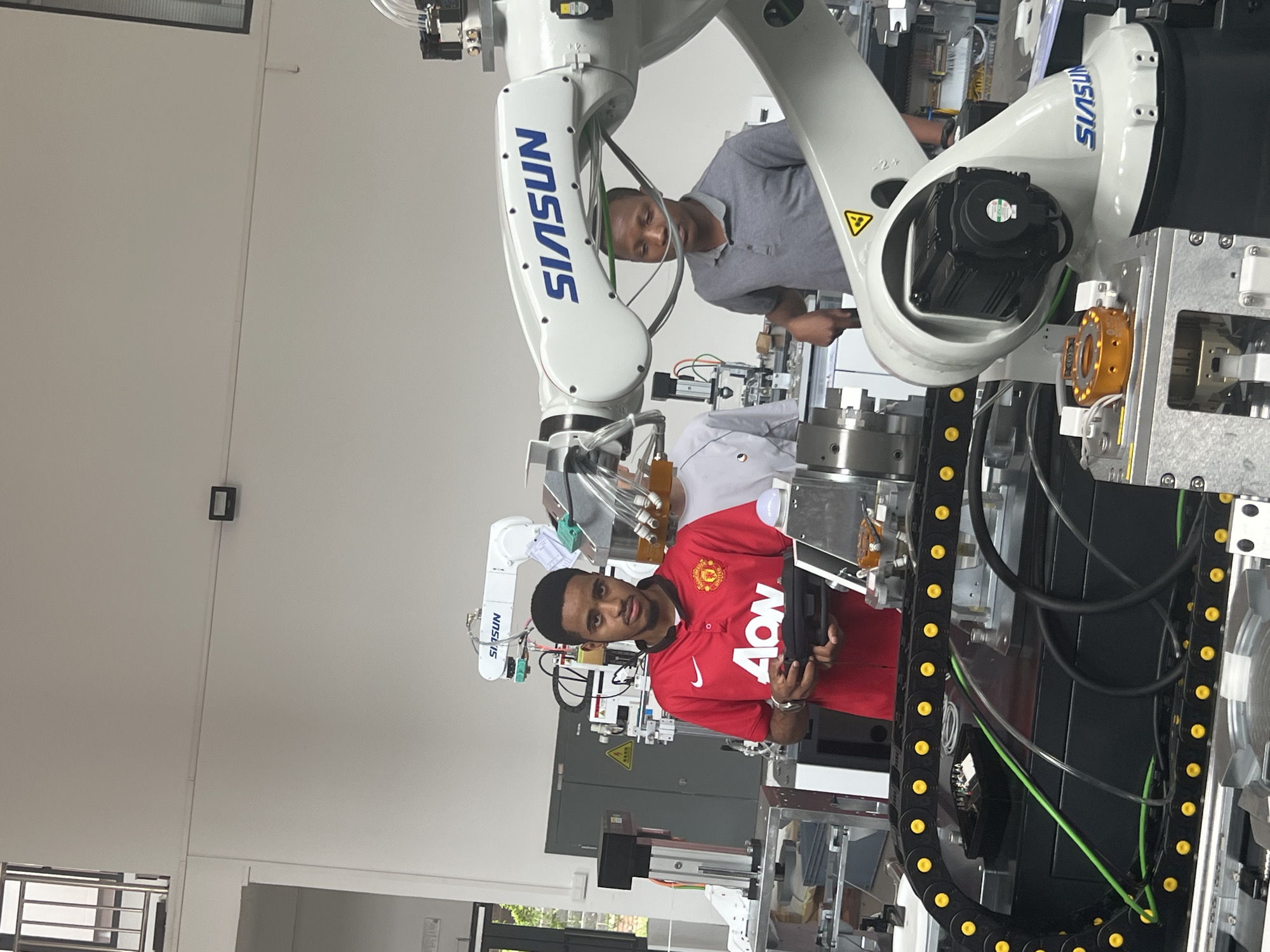 In a well-lit, modern laboratory or workshop environment, two young men are interacting with robotic equipment. The man in the foreground, wearing a red Manchester United jersey with "AON" prominently displayed, is holding a black controller and looking directly at the camera with a neutral expression. Behind him, a robotic arm with the "SIASUN" logo is visible. To the right of the man in red, another man in a grey polo shirt is looking down and appears to be engaged with the machinery. Various pieces of industrial equipment, including a robotic arm, control panels, and metallic structures, are present in the background. The overall setting suggests an educational or research facility focused on robotics or automation. The location is identified as Shuangfu, China.