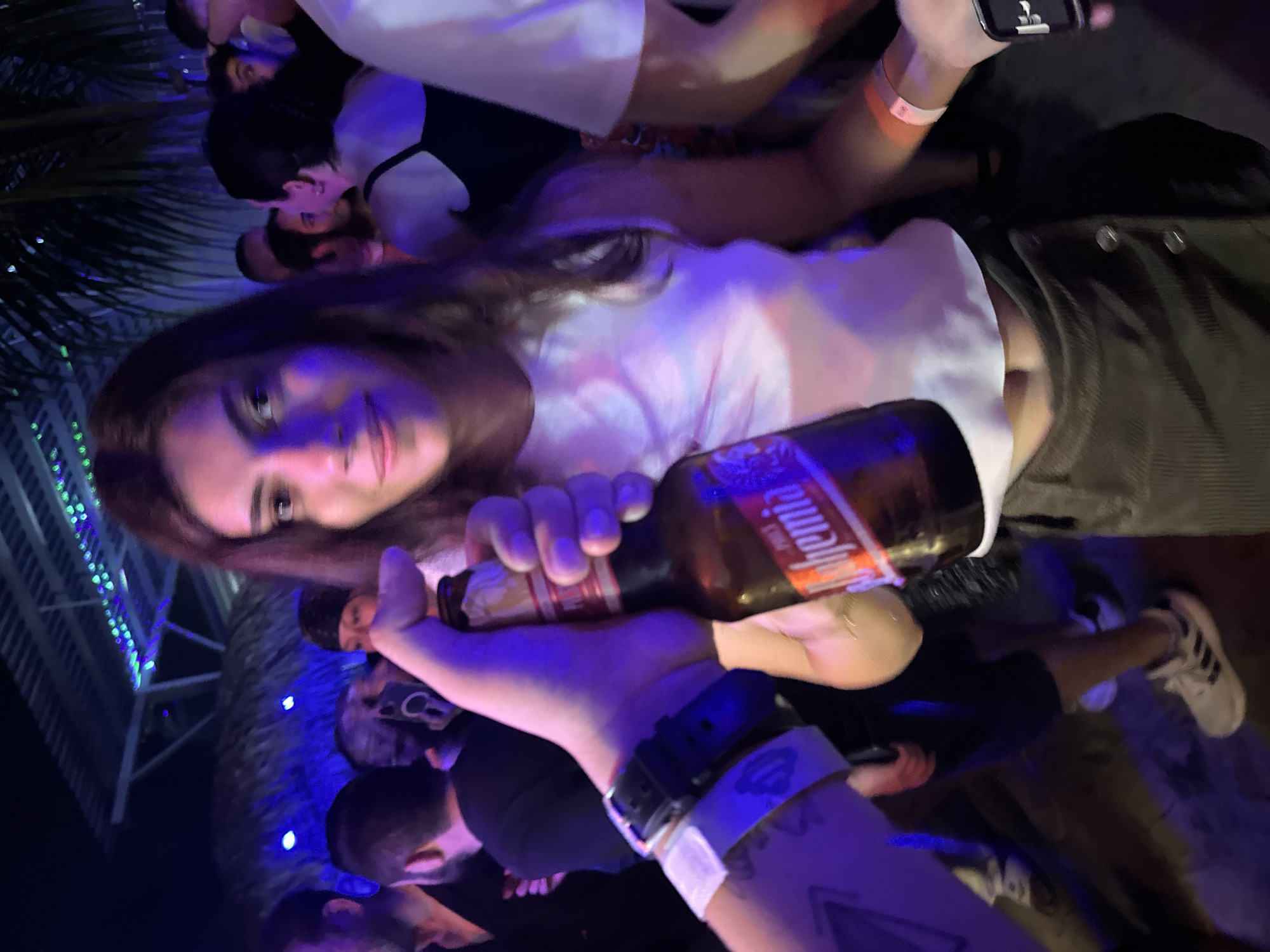 The image shows a group of people in what appears to be a crowded, dimly lit indoor or semi-outdoor setting. A woman with long brown hair is in focus, looking directly at the camera and holding a bottle of "Bohemia" beer. There are other people around her, including a person with visible tattoos on their arm and wrist, holding the bottle close to the camera. Additional individuals are partially visible in the background, suggesting a gathering or party-like atmosphere. Blue lights illuminate the background, and a partial view of what seems to be a palm tree can be seen on the left side of the image. The location is described as Brisas de Zicatela, Mexico, based on external information related to the Bohemia beer brand.