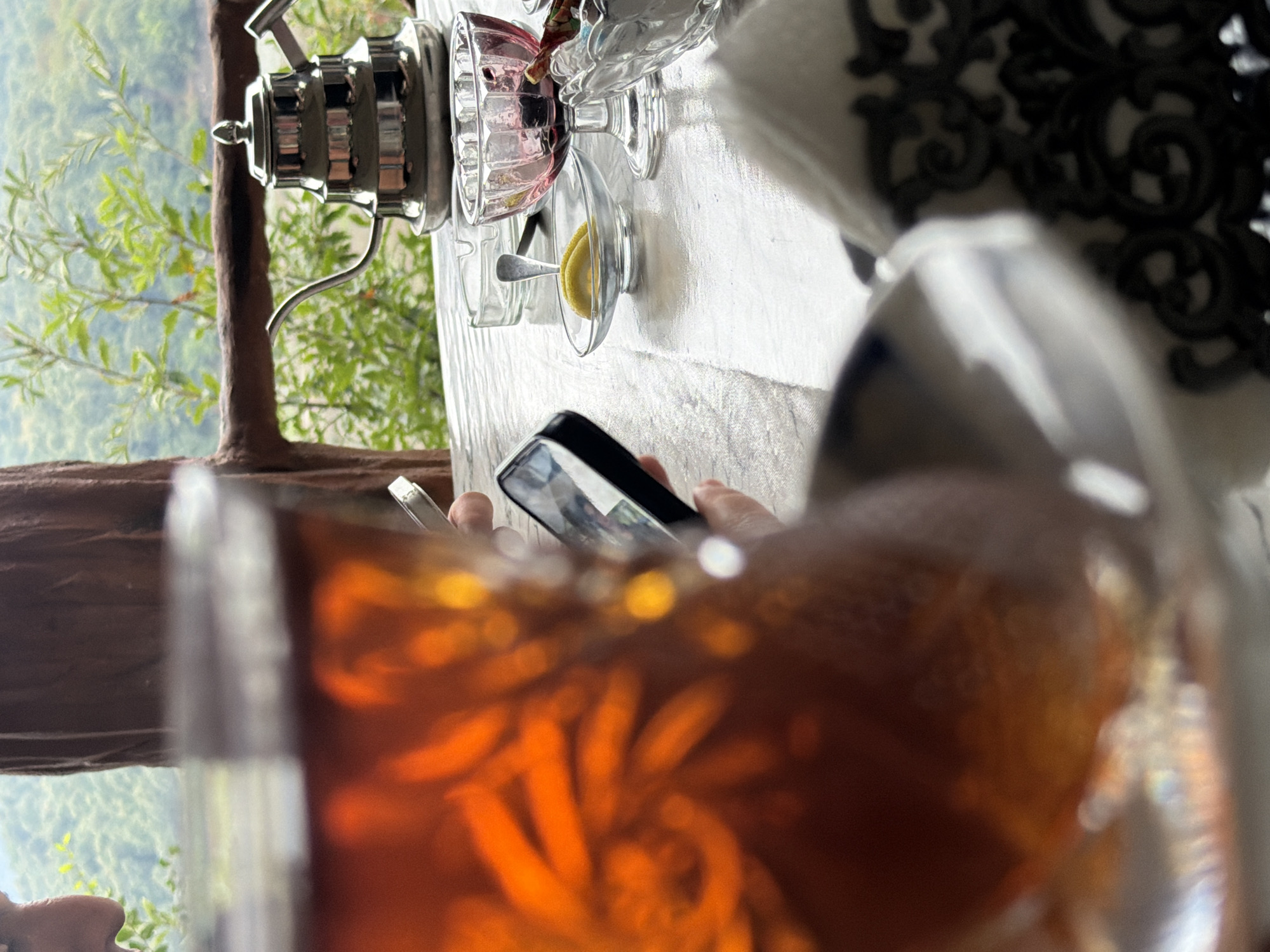 The image depicts a close-up view of a table set for tea with a scenic natural backdrop. In the foreground, a glass of amber-colored tea is prominently featured, its surface reflecting light. To the right, a silver teapot with a distinctive layered design sits on the table. Accompanying the tea are small glass dishes containing what appears to be jam or preserves and a slice of lemon. A person's hand is visible holding a smartphone, suggesting they are engaged with the device. The table is covered with a light-colored tablecloth, and a decorative napkin is placed to the right. In the background, a railing made of natural wood overlooks a lush, green landscape with trees and vegetation, indicating an outdoor setting with a view. The location is identified as Qəriblər, Azerbaijan, suggesting a mountainous or hilly terrain with abundant greenery.