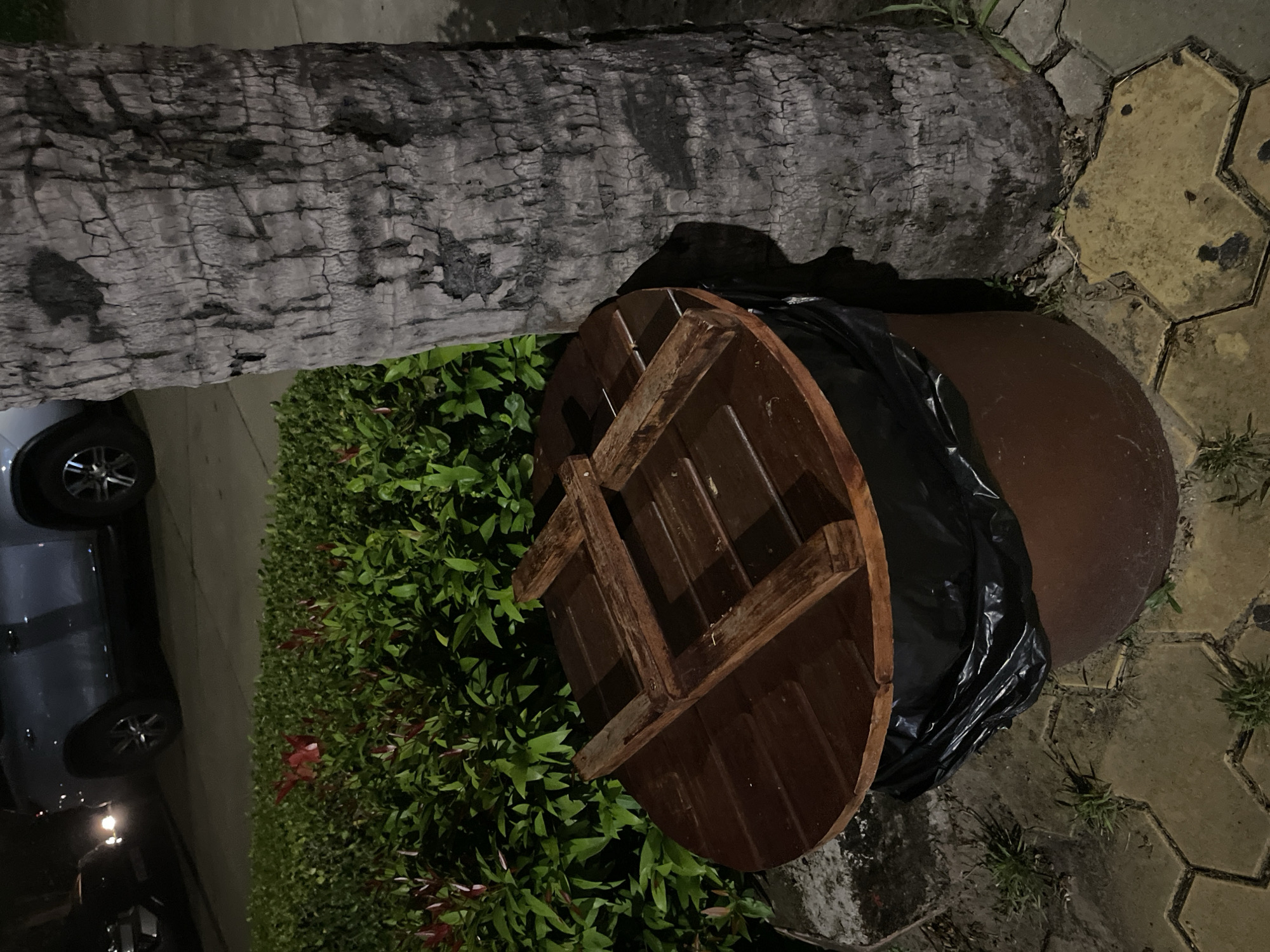The image captures an outdoor scene at night, likely in a public area given the presence of a trash receptacle. The trash can itself is a large, dark brown cylindrical container, with a black trash bag liner visible at the rim. It is topped with a circular, dark wooden lid, featuring a cross-like wooden reinforcement structure secured with screws.
Adjacent to the trash can is a large palm tree trunk, its rough, textured bark predominantly grey. Behind the trash can, a dense green hedge with small red flowers provides a backdrop. In the distance, the headlights of a vehicle are visible, illuminating a portion of the road and the rear of a light-colored SUV. The ground in the foreground is paved with hexagonal yellow and grey interlocking stones, interspersed with patches of short green grass. The overall lighting suggests it is nighttime, with artificial light sources casting shadows. The scene appears to be in a well-maintained urban or suburban outdoor space.