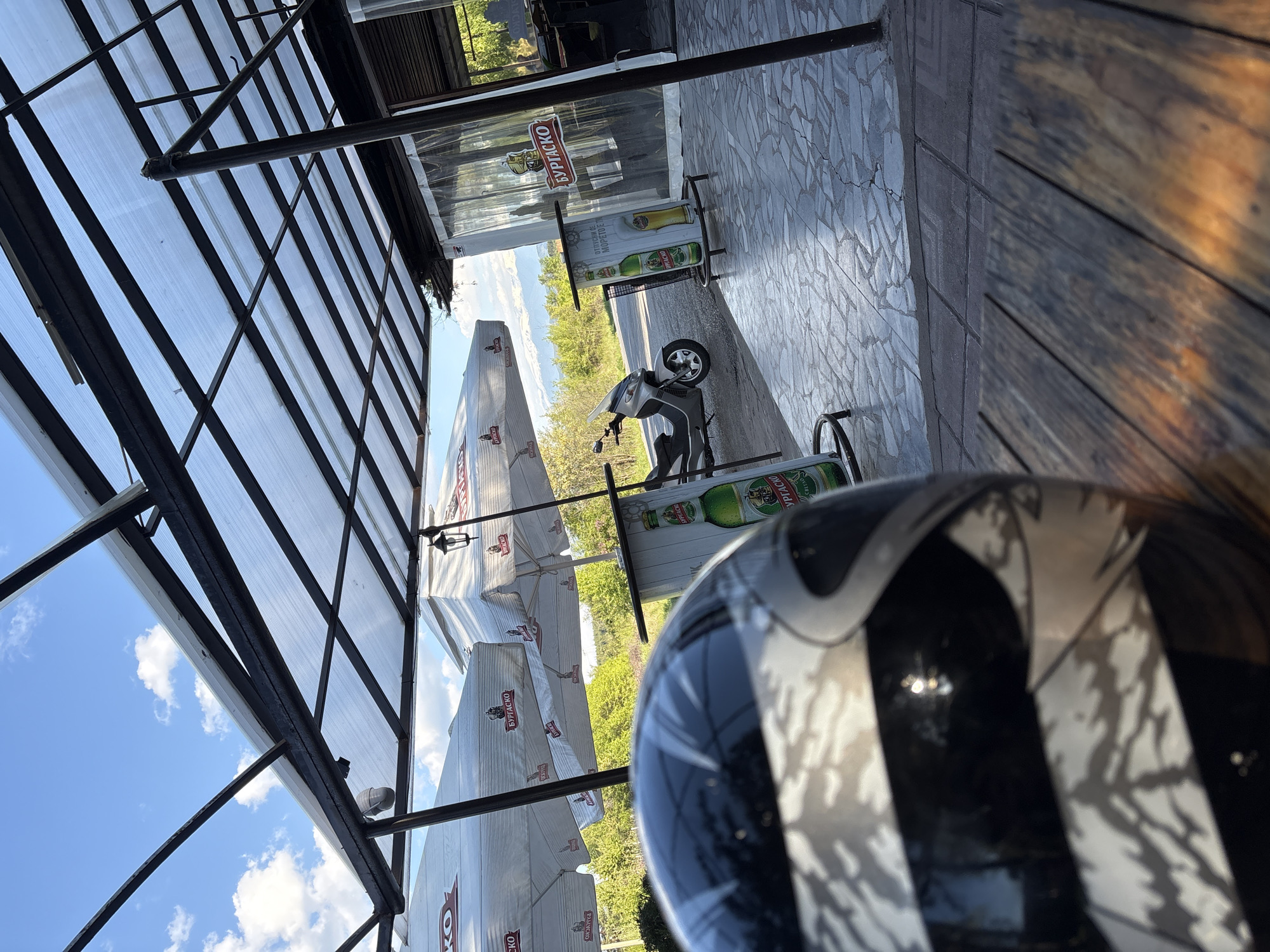 The image shows an outdoor patio scene, likely at a cafe or restaurant in Staro Selo, Bulgaria. A motorcycle is parked on a paved area. Several large umbrellas with the "Burgasko" beer logo are visible. The patio has a covered roof. In the foreground, a motorcycle helmet is partially visible. The weather appears sunny with a blue sky.