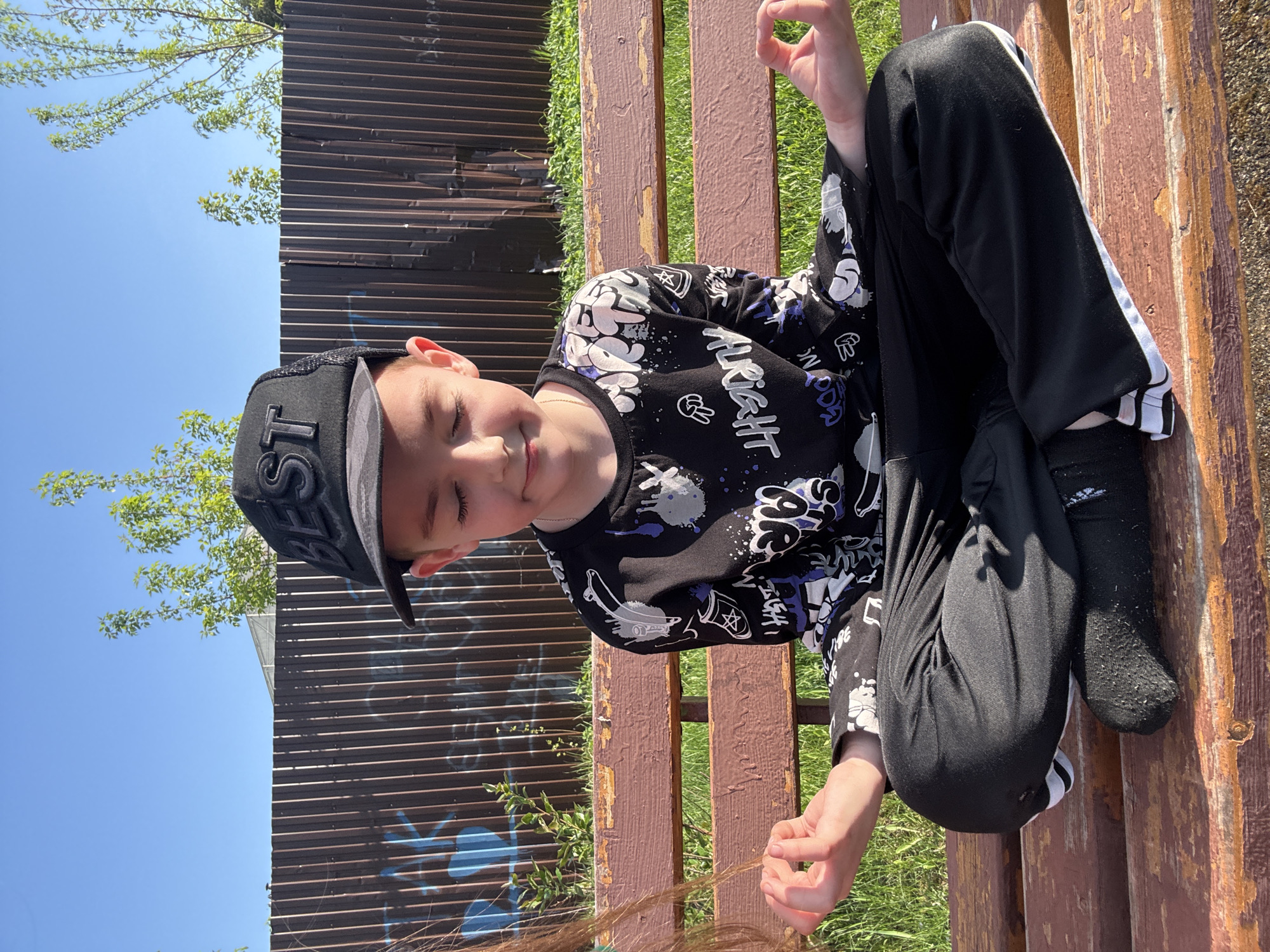 A boy wearing a black baseball cap, patterned long-sleeved shirt, and black pants with white stripes sits on a wooden park bench. The location appears to be outdoors in Раздолье, Russia. A brown fence is visible in the background. The boy is looking down, and part of a person with long brown hair is visible in the lower portion of the image.