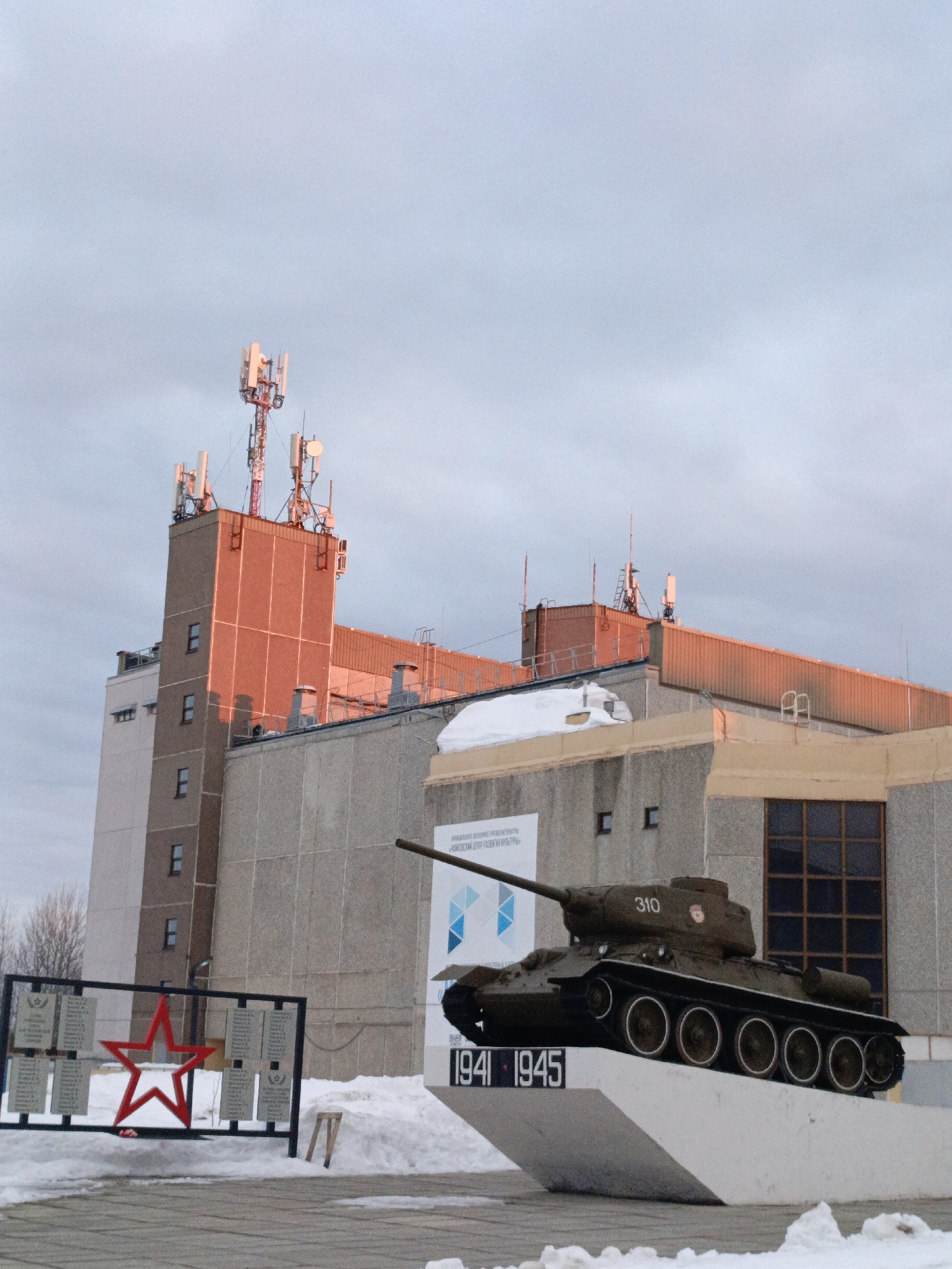 An outdoor scene in the city of Марковский, Russia, depicts a monument featuring a T-34 tank, painted olive green, with the number "310" stenciled in white on its turret. A red Soviet star emblem is visible on the side of the turret. The tank is mounted on a white, angled concrete pedestal, which displays the black numerical text "1941 - 1945" on its front face.
To the left of the tank, a black metal frame holds multiple rectangular memorial plaques, with text in an unidentifiable script, likely Cyrillic. Centered within this frame is a large, three-dimensional red star. Snow covers portions of the ground around the monuments and the base of the buildings.
In the background, a series of multi-story buildings are visible. The leftmost building features a light-colored facade with dark, rectangular windows. Adjacent to it, a taller, reddish-brown building with narrow rectangular windows on its side rises, equipped with multiple cellular or communication antenna arrays on its rooftop. Further to the right, another large, grey concrete building has a large dark-paned window and a banner featuring a blue geometric logo and Russian text. The rooftops of some buildings are covered with patches of snow and reflect a reddish hue from the low-angle sun, contrasting with the overcast, light-blue sky. No people are visible in the scene.