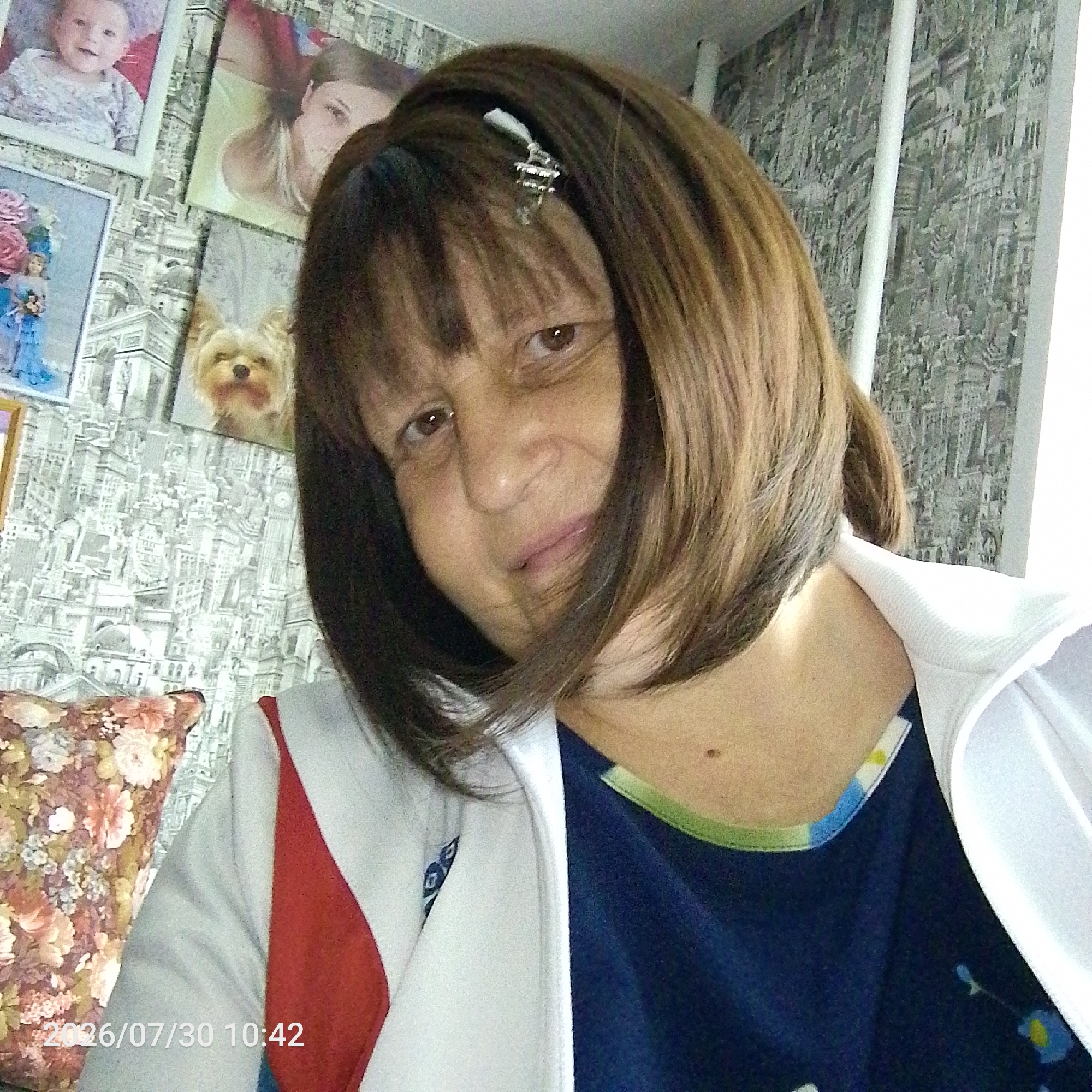 A close-up, waist-up, slightly angled selfie shows a woman with shoulder-length brown hair, bangs, and brown eyes. She is wearing a white jacket over a dark blue shirt with a floral pattern and a green trim around the neckline. A red stripe is visible on the left side of her jacket. In the background, a wall is covered with wallpaper depicting an intricate cityscape with buildings and landmarks. Several framed photographs are mounted on the wall, including one of a baby, a woman, and a dog. A patterned pillow is visible in the lower left corner. The date stamp "2026/07/30 10:42" is overlaid on the image in the lower left corner. The woman is looking towards the camera and has a neutral expression.