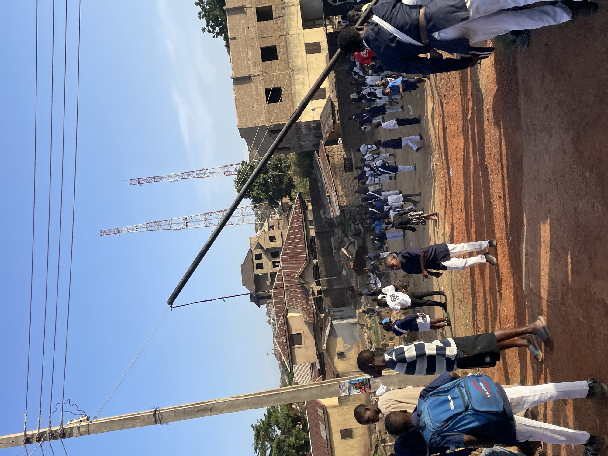 A ground-level, angled shot in Jos, Nigeria shows an outdoor area filled with people, likely school children, dressed in uniforms. The ground is a reddish-brown dirt. People are grouped together, some facing forward, others to the side. In the background are various buildings, some under construction, with exposed brick or concrete. A tall metal pole is visible, extending upwards, with communication towers in the distance against a clear blue sky. Power lines extend from a utility pole. The scene shows an outdoor gathering of students near a building and infrastructure.