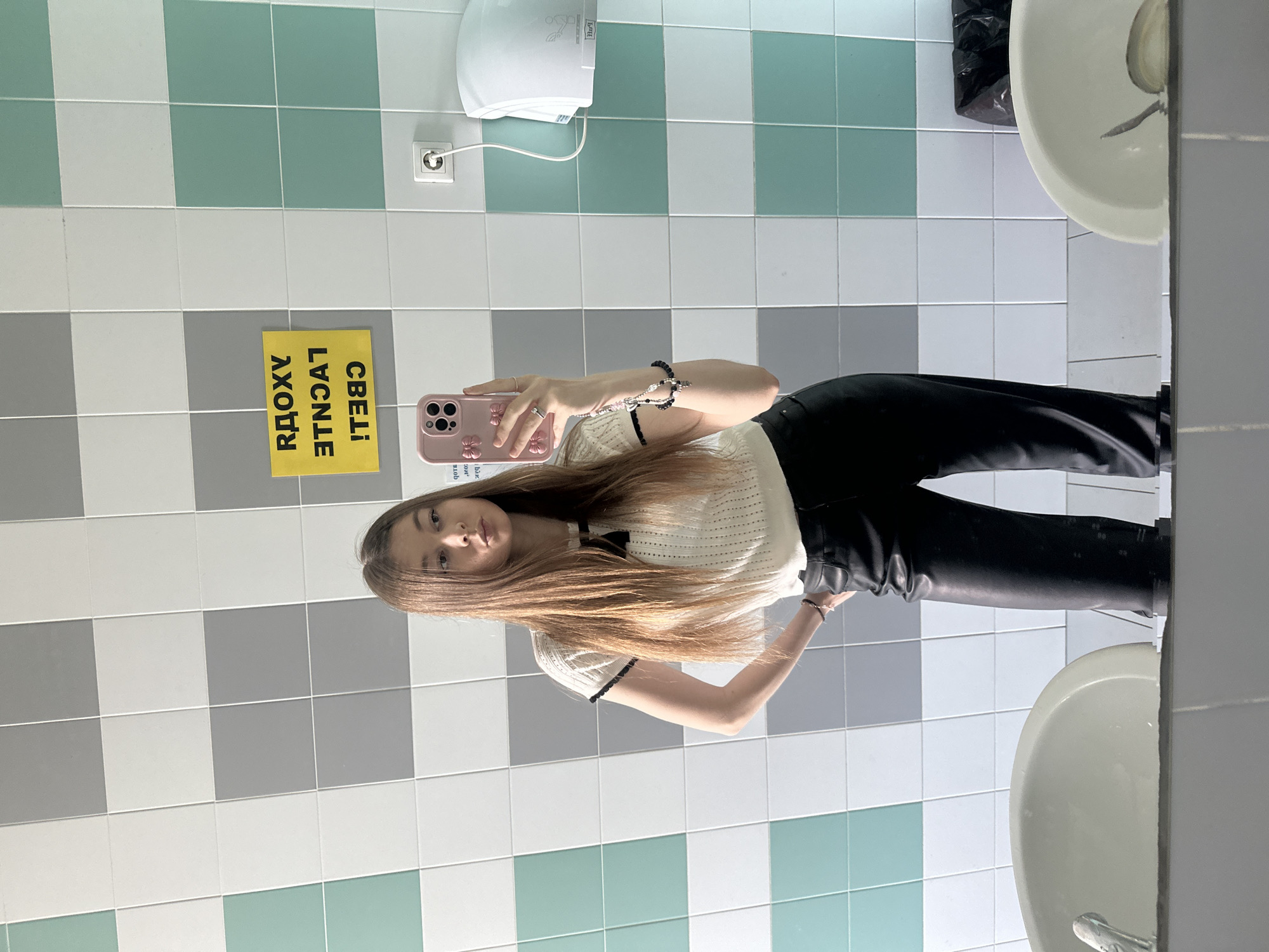 A young woman is taking a selfie in a public restroom. She is holding a pink cell phone with a floral design on the case. The woman is wearing a light-colored, textured top with black trim and dark pants. Her hair is light brown and long. The background consists of tiled walls with a pattern of alternating green, white, and gray squares. A hand dryer is mounted on the wall. A yellow sign with reversed Cyrillic text is affixed to the wall. Two white sinks are visible. The location is likely in the city of Kazan, Russia, based on the Cyrillic text on the sign.