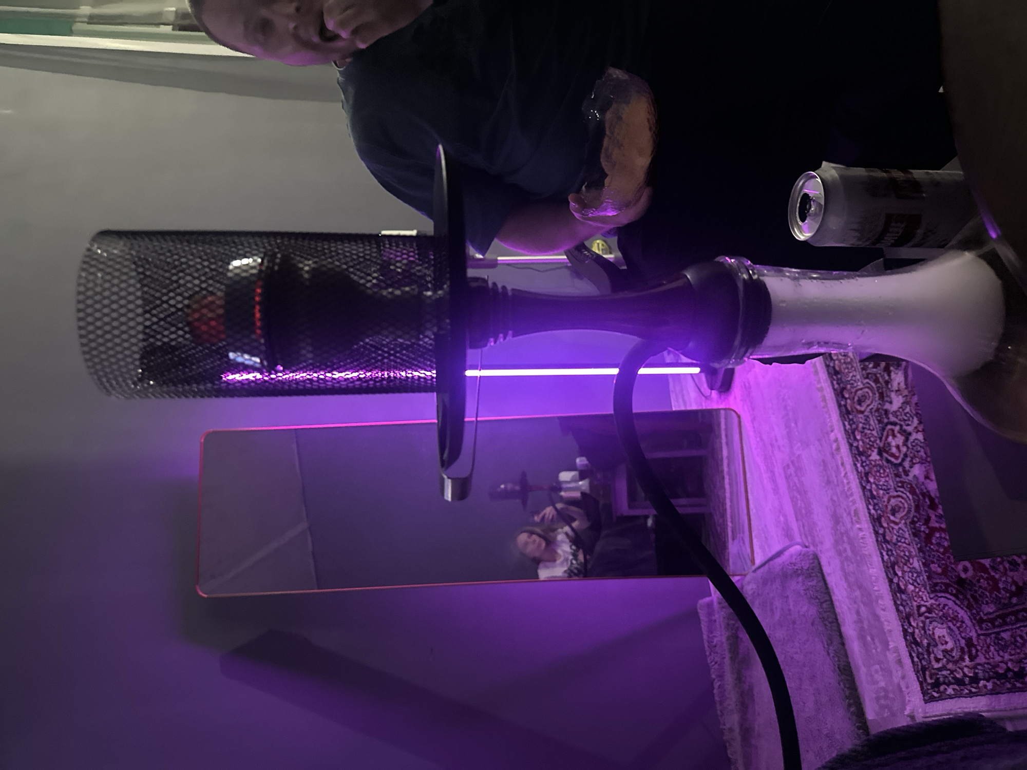 The scene depicts a person holding a hookah, which is emitting smoke. A can of beer sits nearby. A mirror reflects another person in the background. The room is illuminated with purple lighting. The presence of a patterned rug and the hookah suggests a recreational or social setting. It is not possible to definitively establish that the image was taken in Blagoveshchensk, Russia from the content given.