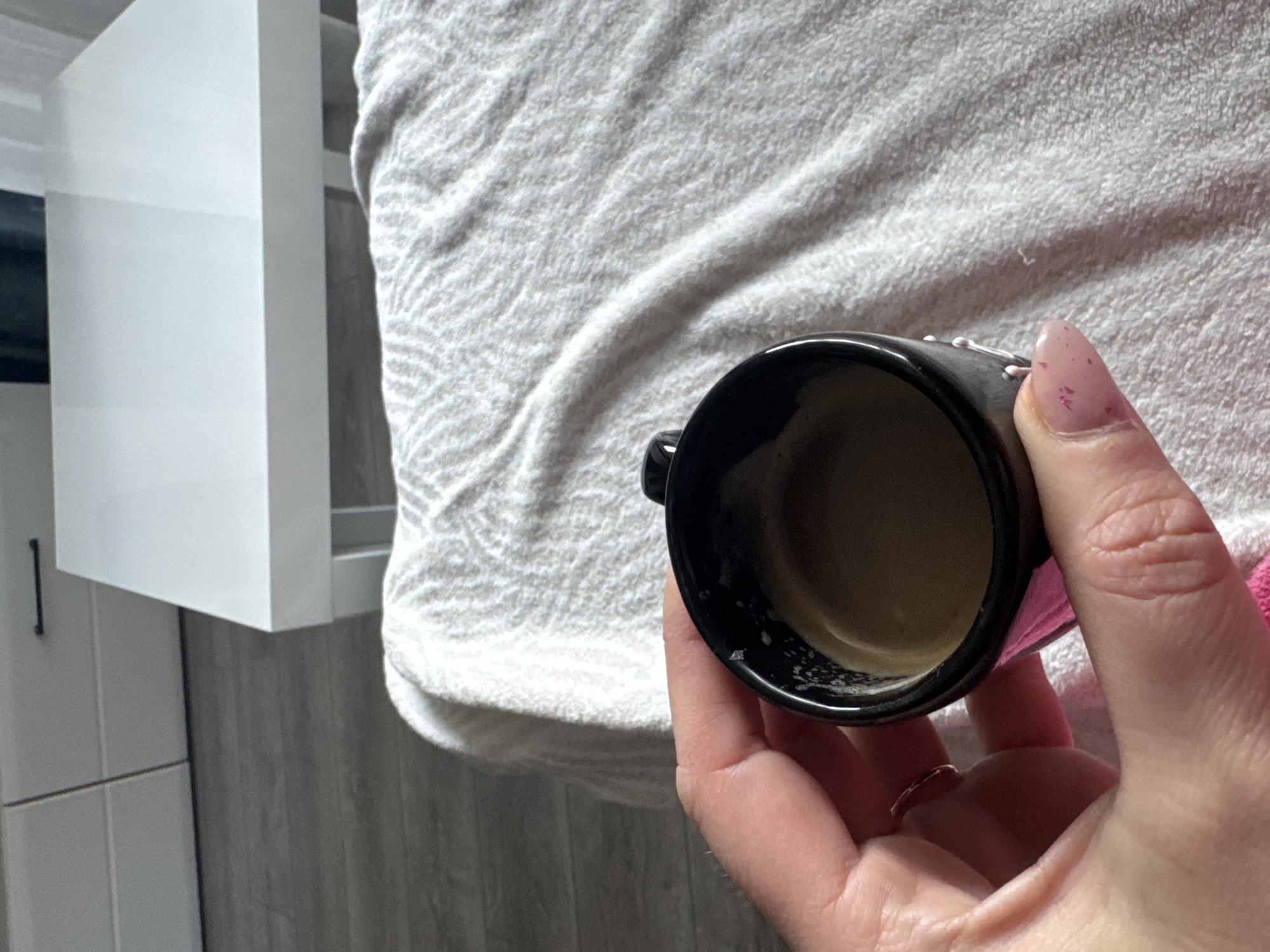 A hand holds a small black cup filled with light brown liquid. The background includes a grey and white textured blanket, a white cabinet, and a grey floor. The location is not discernible from the image.