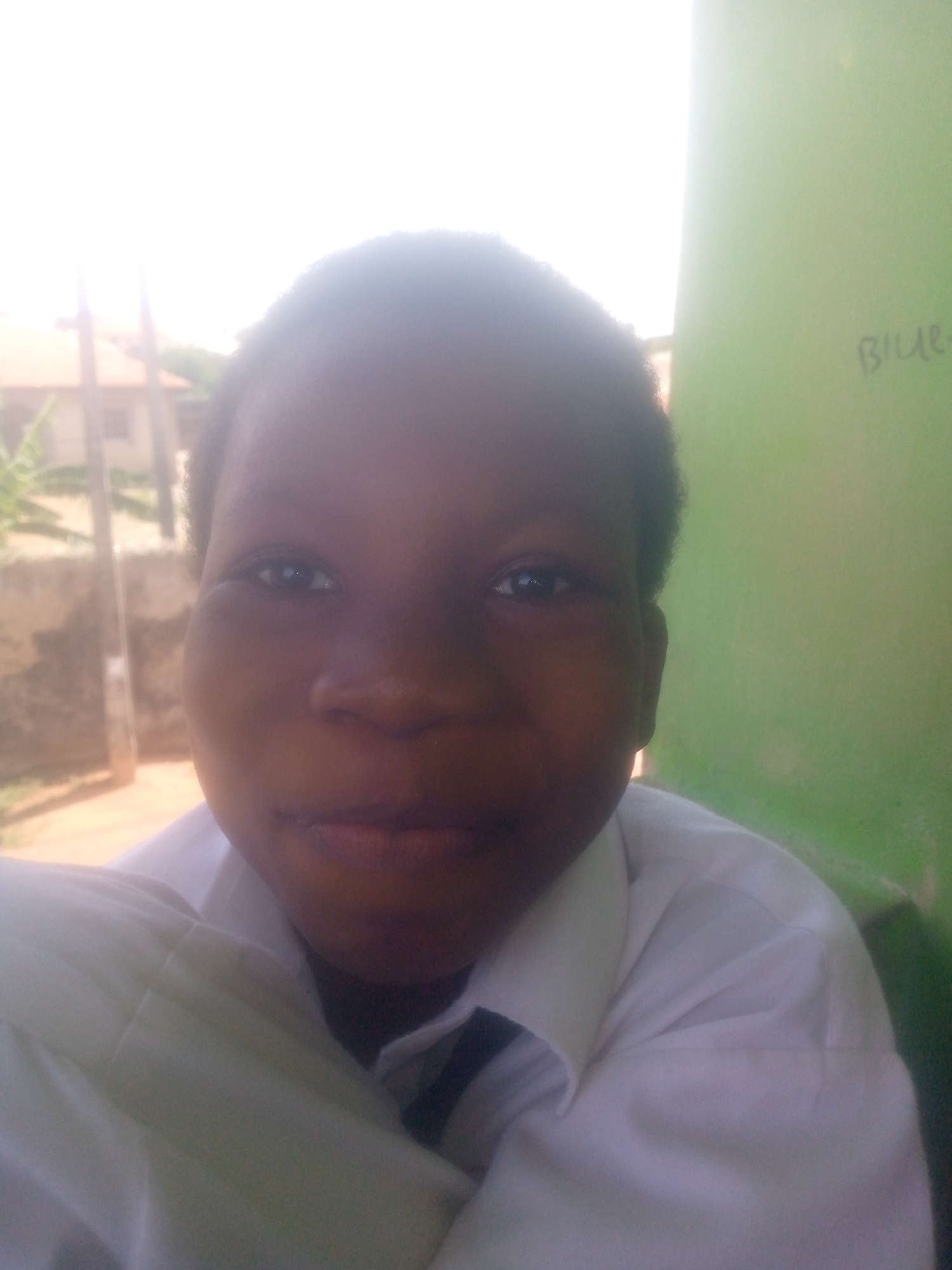 A close-up shot of a young person with dark skin, wearing a white collared shirt, likely a school uniform. The background is slightly blurred, but reveals a building exterior on the left and a green-painted wall on the right, with the word "BLUE" faintly visible. The location appears to be outdoors during daylight hours.
