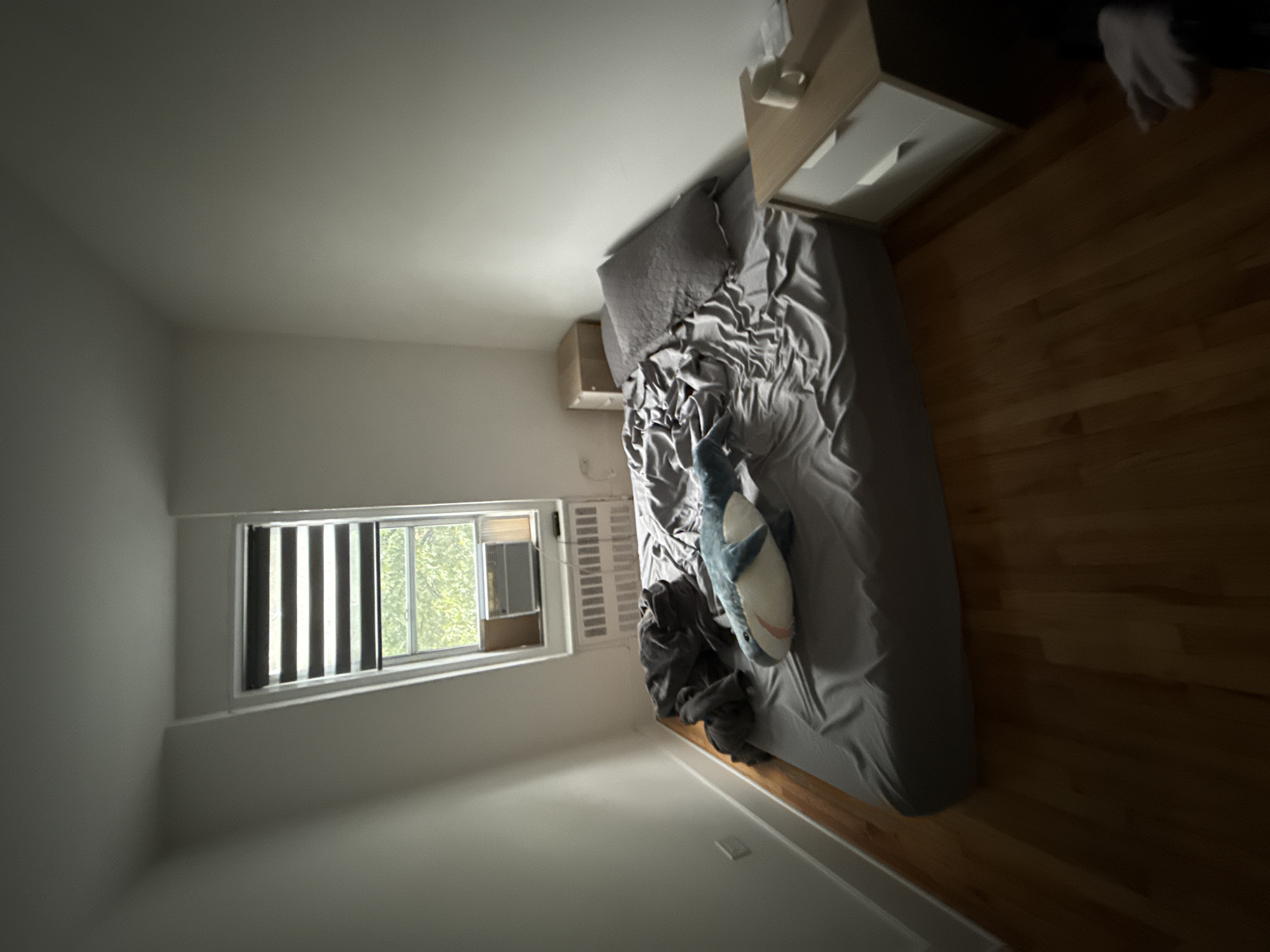 The image depicts a bedroom in the City of Yonkers, United States of America. A mattress, unmade with gray bedding, dominates the center of the room, with a plush shark toy resting on it. To the left of the bed, a pile of dark gray clothing is visible. A window with horizontal blinds is positioned on the far wall, above a white radiator and an air conditioning unit. Adjacent to the window, a small, light brown nightstand is placed. To the right of the bed, a white nightstand with two drawers is situated. On this nightstand, a white mug and some papers are present. The room has light-colored walls and a polished wooden floor. The lighting suggests it is daytime, with natural light filtering through the window, casting shadows across the room.