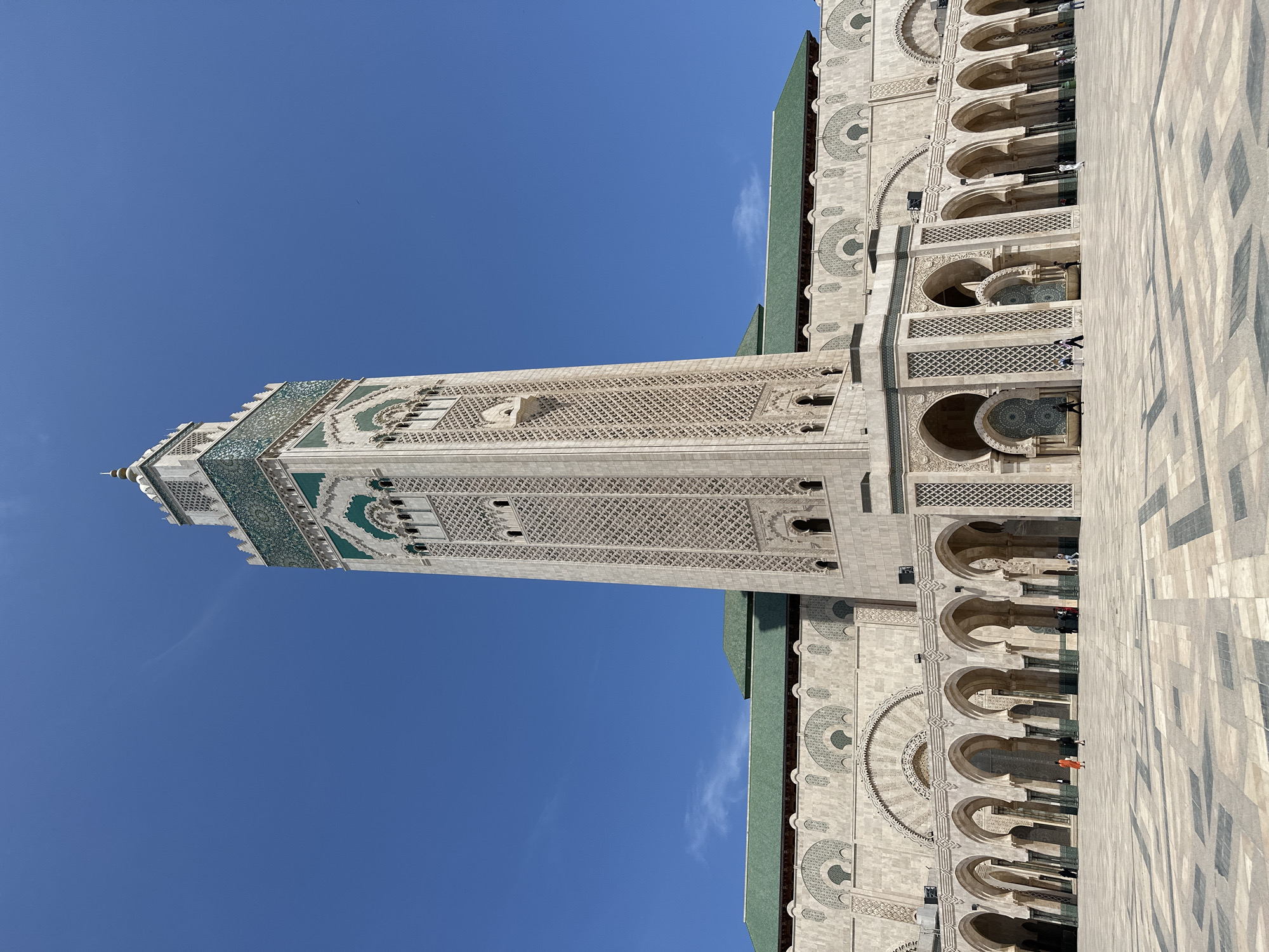 The image depicts a majestic architectural scene, showcasing the impressive Hassan II Mosque and its towering minaret in Casablanca, Morocco, under a clear blue sky. The main subject is the mosque's minaret, which stands tall on the left side of the frame, constructed from light-colored stone and intricately adorned with geometric patterns and sections of green and white mosaic tiles (zellij). The minaret tapers slightly towards its pinnacle, which features a pointed finial.
To the right of the minaret, the main body of the mosque is visible, also made of light-colored stone with complex decorative carvings and large arched windows or entrances. These arches are symmetrically arranged and feature detailed stonework. A distinct green-tiled roof is visible atop parts of the mosque structure.
In the foreground and extending to the right, a vast, patterned paved courtyard stretches out. The paving consists of light and darker rectangular tiles arranged in a geometric design. Scattered across this expansive courtyard and near the mosque's entrances are several people, appearing relatively small against the monumental scale of the architecture. They are mostly seen walking or standing, suggesting a general visitation or public access to the mosque grounds.
The scene is set outdoors on a bright, sunny day, indicated by the brilliant blue sky and the distinct shadows cast by the mosque and individuals on the courtyard pavement. The overall impression is one of grandeur, intricate craftsmanship, and serenity. There is no visible text in the image.