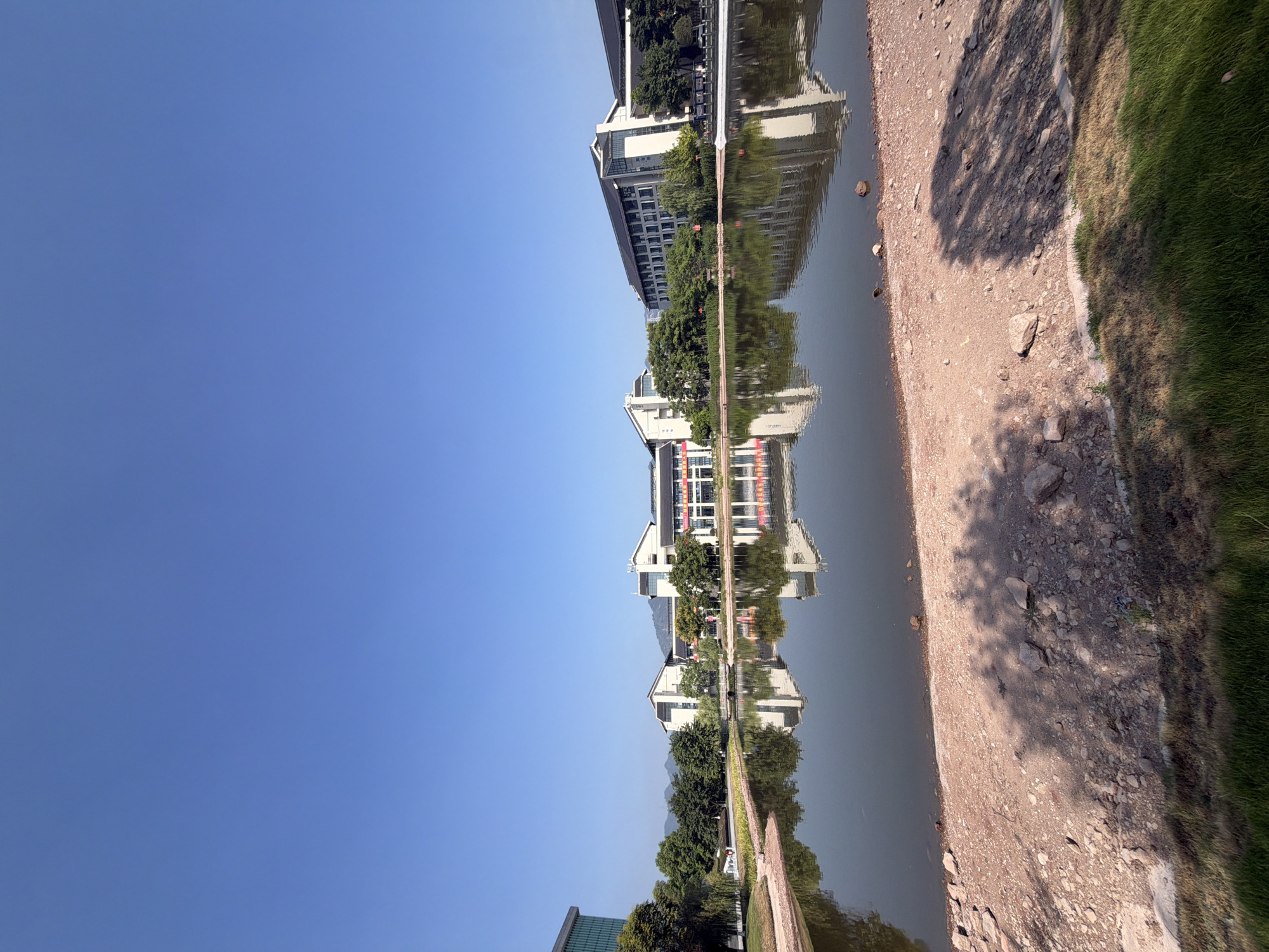 The image shows a serene waterscape with modern buildings reflected in the calm water of a lake. The foreground features a dry, sandy embankment with scattered rocks and patches of grass. The lake, with its smooth surface, mirrors the clear blue sky and the architecture of the buildings lining its opposite shore. These buildings are a blend of contemporary design with traditional Chinese architectural elements, characterized by dark tiled roofs and white facades. Lush green trees are interspersed between the structures, adding to the picturesque setting. The overall atmosphere is peaceful and idyllic. The location is identified as the campus of Chongqing University of Technology, Jiangjin District, China. There are no people or animals visible in the image.