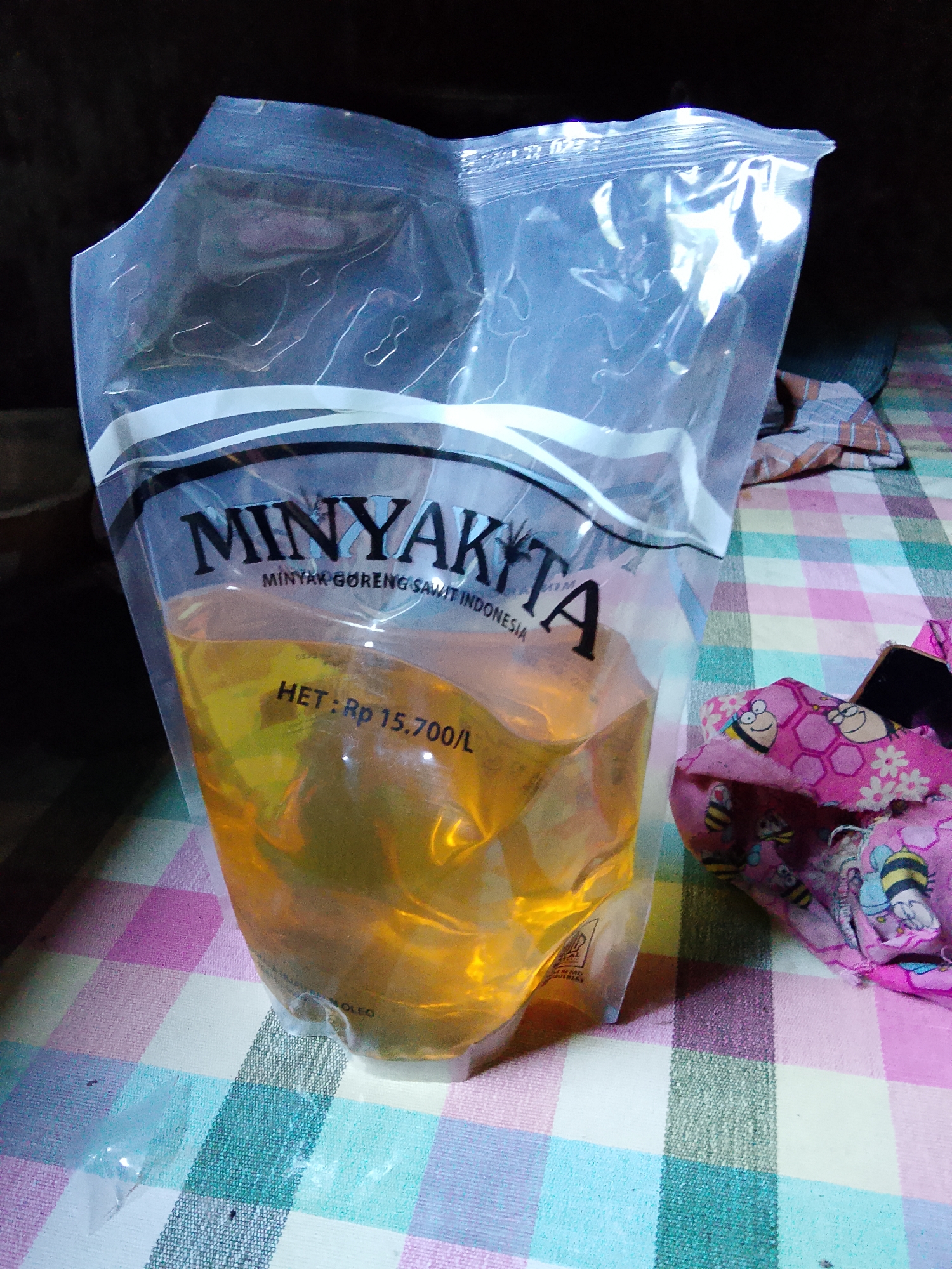 An unsealed, transparent plastic pouch containing golden cooking oil is positioned on a table covered with a checkered tablecloth. The pouch displays the brand name "MINYAKITA" in bold, dark lettering with "MINYAK GORENG SAWIT INDONESIA" printed below it in a smaller font. The price "HET : Rp 15.700/L" is also visible on the pouch. To the right of the pouch, a pink fabric with a pattern of bees and flowers is visible, along with a dark object, possibly a smartphone. The background is dark and out of focus. The image appears to have been taken indoors. There is no indication of the location in the city of Merbopuntong, Indonesia.
