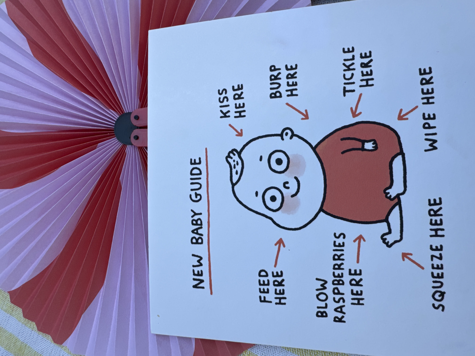 The image displays a handmade card with the title "NEW BABY GUIDE". The card features a cartoon drawing of a baby with various instructions pointing to different parts of its body. These instructions include "FEED HERE", "BLOW RASPBERRIES HERE", "SQUEEZE HERE", "KISS HERE", "BURP HERE", "TICKLE HERE", and "WIPE HERE". The card is placed on a yellow and white striped surface, and a pink and red folded fan is visible in the background. The scene is indoors, with soft lighting. There are no people present in the image, and the focus is solely on the card and the fan. The image was taken from a slightly elevated angle, looking down at the card. The location in Agios Tychona Community, Cyprus, is not depicted in this image; the image shows a close-up of a card with a drawing of a baby.