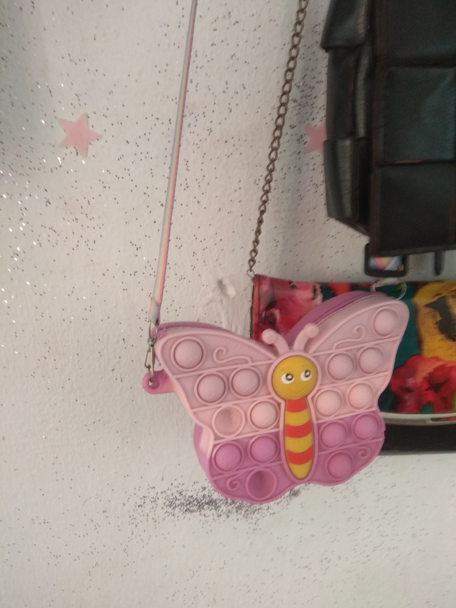 A pink butterfly-shaped fidget toy purse hangs from a metal chain against a white, textured wall. The wall is speckled with tiny, dark dots and scattered glitter. To the left, a pale pink star sticker is visible. The butterfly purse features a yellow face with white eyes, a red and yellow striped body, and a series of circular depressions on its wings, characteristic of a fidget toy. A colorful strap, striped with pink, yellow, and blue, is attached to the purse. Behind the purse, parts of other patterned items are visible, including a floral print and a vibrant abstract design. To the right, a dark, textured object with a grid-like pattern is partially visible, possibly a bag or a piece of furniture. The lighting suggests it is daytime, and the overall scene appears to be indoors, possibly a child's room given the playful nature of the purse and wall decorations. There is no discernible text in the image.