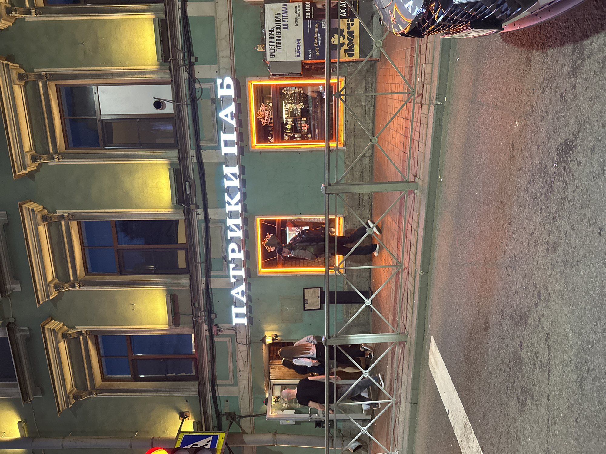 The image captures a street scene at night in Saint Petersburg, Russia, in front of a establishment with a neon sign that reads "ПАТРИКИ ПАБ" (Patriki Pub). The building facade is painted a muted green and features several windows with ornate moldings. Warm yellow light emanates from the windows and a neon orange outline frames one of them, which displays a bar interior with bottles of alcohol. A metal railing with a diamond pattern separates the sidewalk from the street. Several people are visible: two men and two women are walking towards the entrance of the pub, with one man and one woman already entering. Another man with a backpack is walking away from the entrance. A traffic light with a red light illuminated is visible on the left side of the frame, along with a traffic sign. A parked car is partially visible on the right side of the image. The sidewalk is paved with reddish-brown bricks, and the street is asphalt. Advertising posters are displayed on the wall of the building.