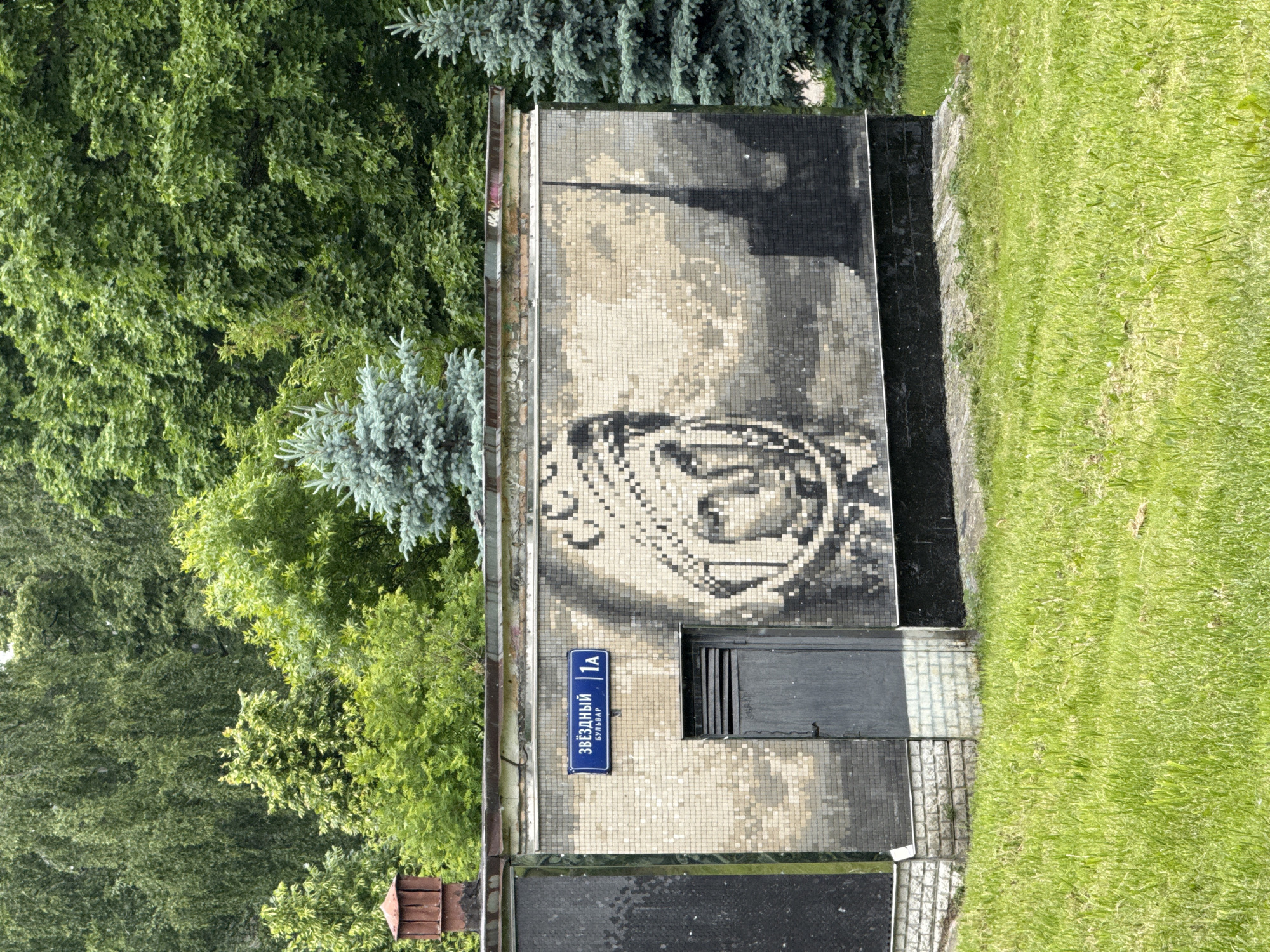 The image captures an outdoor scene featuring a building wall adorned with a large mosaic, surrounded by lush vegetation. The wall, positioned centrally in the frame, is covered in a mosaic composed of square tiles in shades of white, beige, grey, and black. The artwork depicts a stylized human head or face, possibly in profile, within a circular or emblem-like design, suggesting a monumental or historical theme.
Below the mosaic, a dark, rectangular door or vent opening is visible. A blue street sign is affixed to the wall, to the left of this opening. The lower section of the wall features a dark, reflective panel at its base, next to a small paved area with tiles and vibrant green grass.
Dense, verdant trees, including both broadleaf and coniferous varieties (like blue spruces), fill the left and top portions of the image, indicating a green, possibly park-like setting. Bright green grass extends across the bottom and far right of the frame. The scene is brightly lit by daylight, suggesting clear weather.
The blue street sign clearly displays the Cyrillic text: "ЗВЁЗДНЫЙ" (Zvyozdnyy) at the top, "БУЛЬВАР" (Bulvar) below it, and "1А" to the right. This translates to "Starry Boulevard, 1A," a location in Moscow, Russia. No individuals or specific activities are depicted.