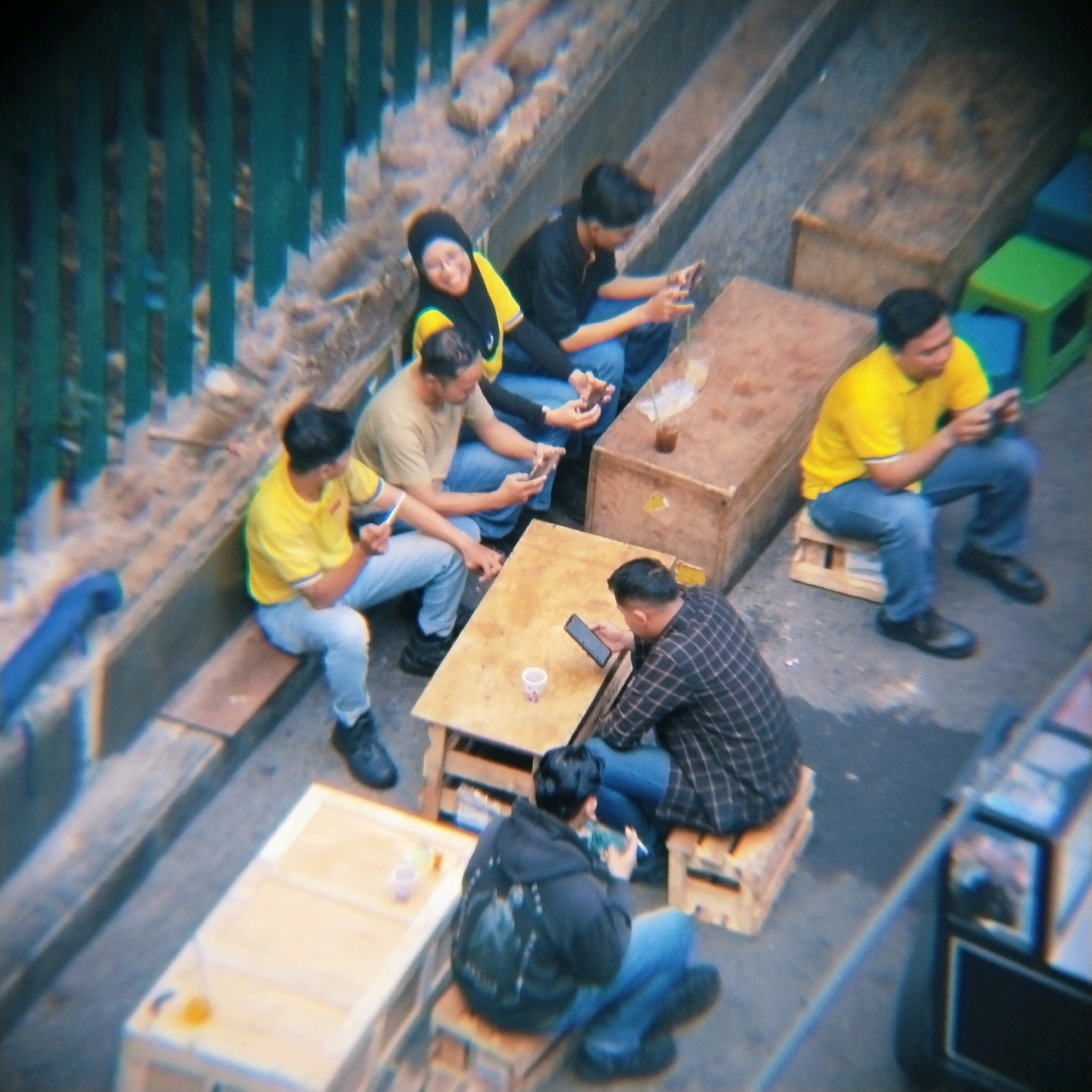 The image depicts an outdoor setting with several individuals gathered around low tables. The environment suggests a casual, possibly street-side seating area in Surabaya, Indonesia, indicated by the presence of what appear to be makeshift wooden tables and stools, and a green and blue plastic stool commonly found in such informal establishments.
On the left, two men are seated on the ground. The man in the foreground, wearing a light-colored t-shirt and blue jeans, is looking down at his phone. To his left, another man in a yellow t-shirt and blue jeans is also focused on his phone. Adjacent to him, a woman wearing a yellow and black headscarf and black jacket is visible, smiling and looking towards the viewer. She is seated next to a man in a black t-shirt and blue jeans, who is also engrossed in his phone.
In the center of the image, a wooden table is positioned between two men. One man, wearing a light brown t-shirt and blue jeans, is looking at his phone. The other man, in a black and white checkered shirt and blue jeans, is also looking at his phone. A small cup is on this table.
Further to the right, a man in a bright yellow polo shirt and blue jeans is seated on a wooden stool, holding a phone. Behind him, a large wooden crate serves as a table. Another wooden crate is visible in the background, with two green plastic stools and a blue plastic stool.
The overall scene portrays a group of people enjoying a casual gathering, possibly at a street food stall or small eatery, where mobile phone usage is prevalent. The lighting suggests it is daytime.