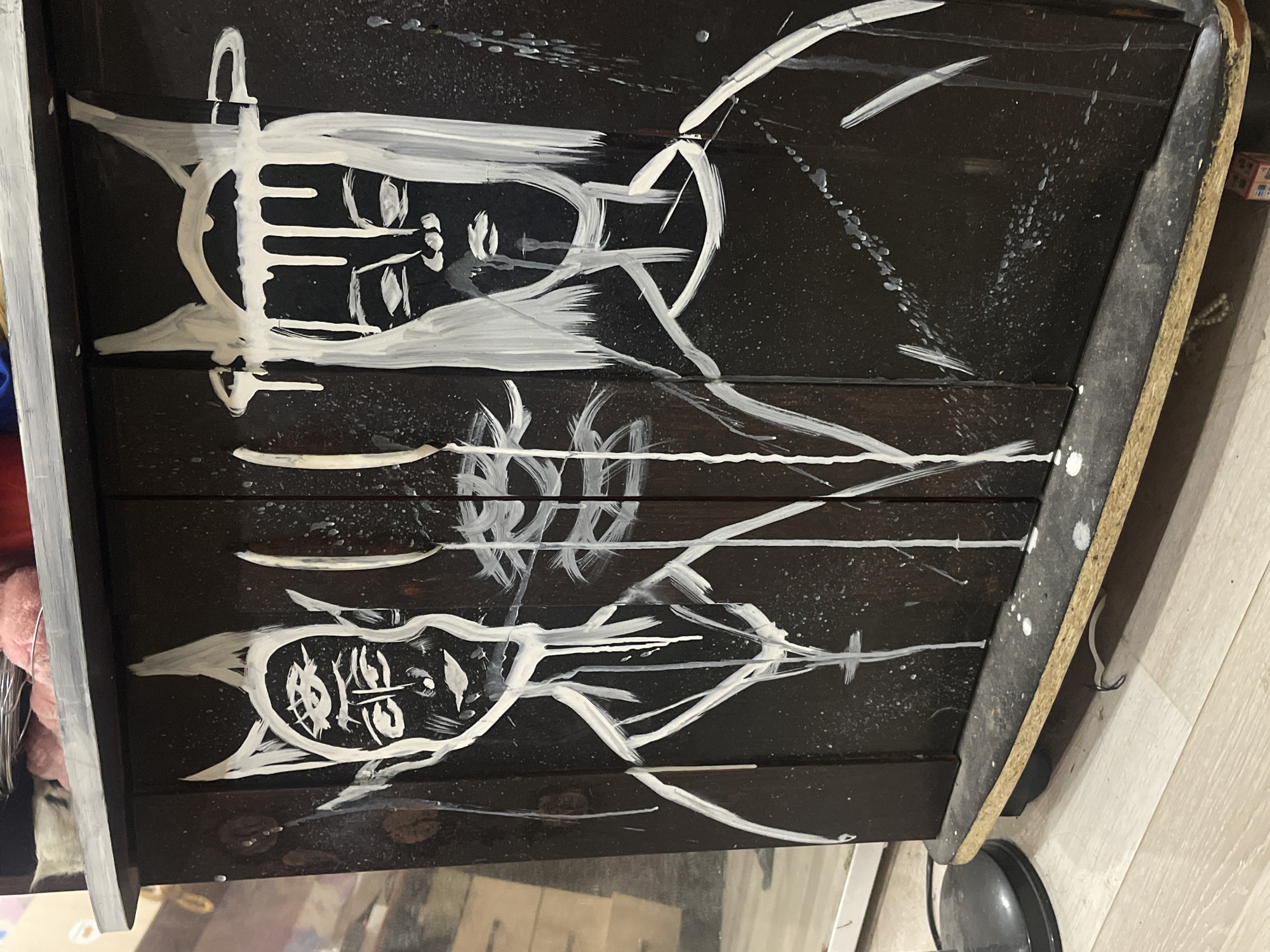 The image shows the front of a dark wooden cabinet with two stylized figures painted in white. The figures are abstract and have a somewhat unsettling, almost skeletal appearance. The figure on the left has pointed, horn-like ears and multiple eyes. The figure on the right has a dripping crown-like headdress. Both figures have stark, minimalist facial features and their bodies are outlined with thin white lines, with some paint splatters and drips on the surface of the cabinet. The cabinet appears to be a piece of furniture, possibly a cabinet or a chest of drawers, with a wooden base. The background is blurred but suggests an indoor environment with some objects and furniture. There is no specific indication of the location in the city of ЖК «Борисоглебское», Russia.