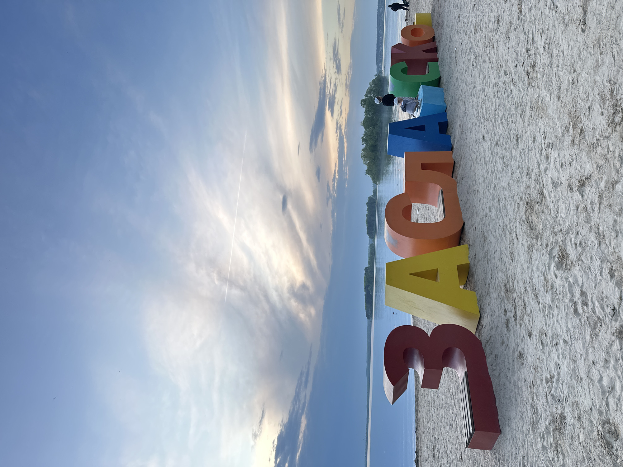 The image captures a landscape scene, likely taken in or near Ждановіцкі сельскі Савет, Belarus, given the information provided. Dominating the foreground are large, colorful three-dimensional letters spelling out a word in Cyrillic. The letters, which appear to be made of a solid material, are positioned on a sandy surface, possibly a beach.
In the background, a body of water is visible, possibly a lake or river. Beyond the water, a strip of green vegetation, likely trees, forms a horizon line. Above the trees, the sky is visible, with streaks of clouds.
The scene appears to be lit by natural sunlight, as indicated by the shadows and overall brightness of the image.
Two people are on the top of one of the letters. Another person is visible in the distance, near the end of the letters.