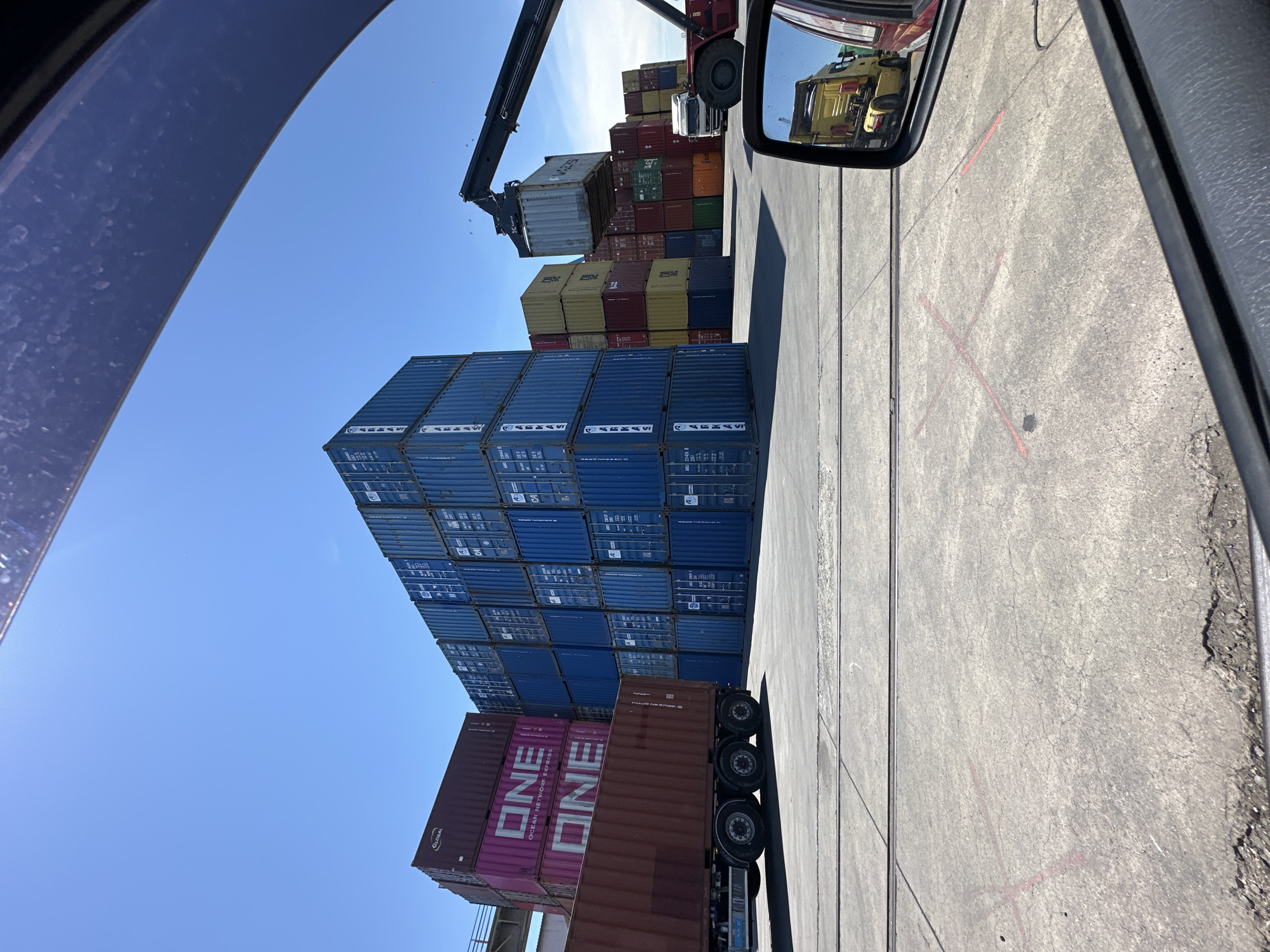 The image depicts a sunny day at a shipping container yard. In the foreground, a portion of a vehicle's interior is visible, including a side mirror reflecting a yellow truck. The ground is paved with concrete, marked with red lines forming an 'X' and a shorter line. Several stacks of shipping containers dominate the scene. On the left, a maroon truck is parked, loaded with a maroon shipping container. Next to it, a stack of pink and maroon containers, some branded with "ONE," are also visible. Further back, tall stacks of blue containers form a wall on the right. In the middle ground, a large forklift or crane is in operation, lifting a container. Behind these, more stacks of containers in various colors like yellow, red, green, and blue are arranged. The sky is a clear, bright blue.