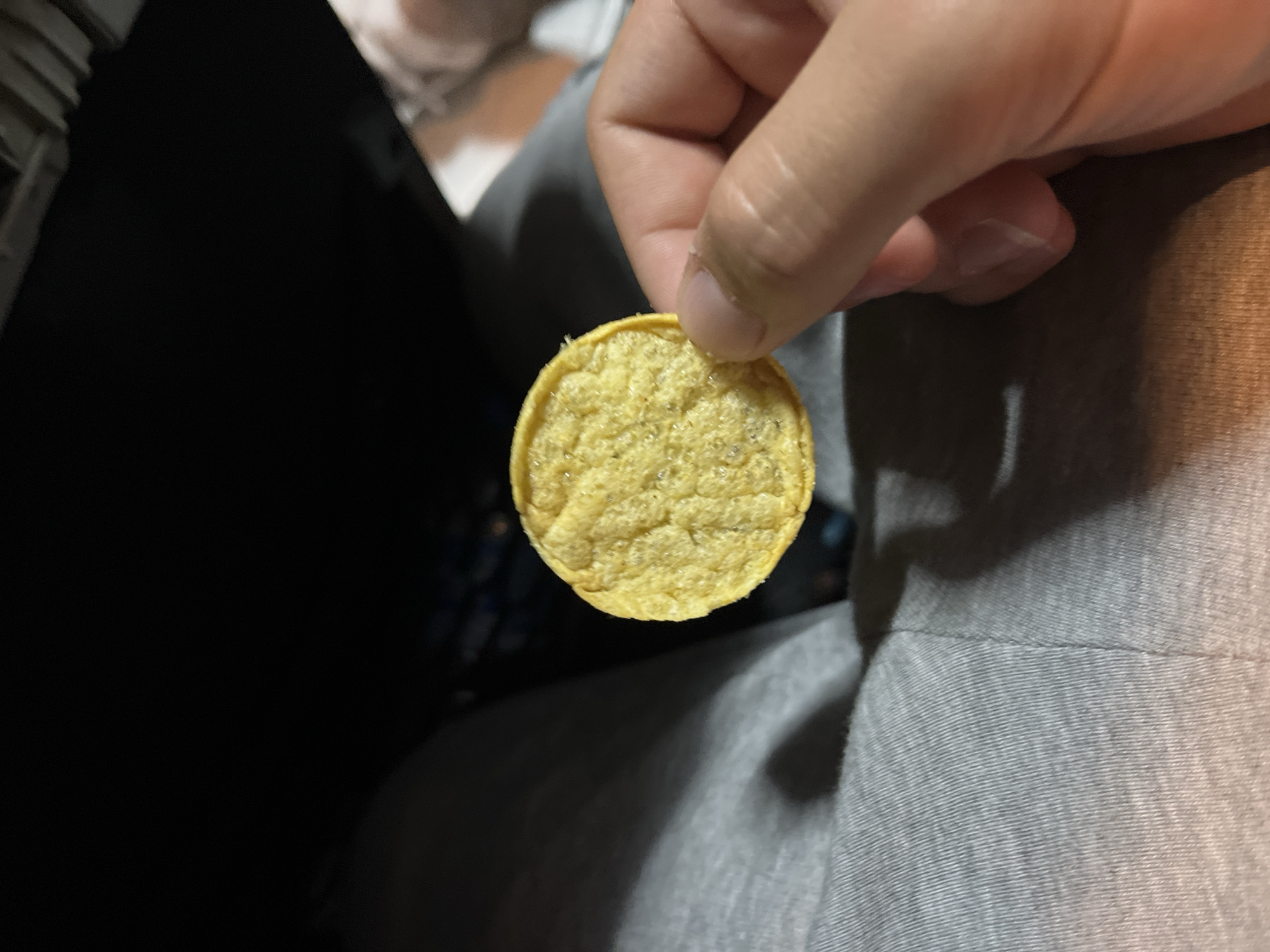 A hand holds a single, round, yellow potato chip with a textured surface. The background consists of a dark area and a grey surface, potentially clothing. The image does not contain information about Popovitsa, Bulgaria.