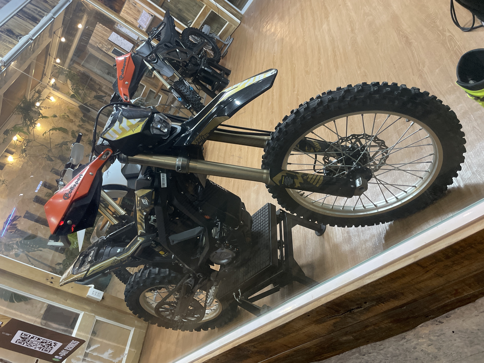 The image shows two dirt bikes inside a store. The bike in the foreground is black and gold with orange handguards and a yellow and black graphic on the front fender. It is propped up on a black stand. The second dirt bike, visible in the background, is also black and appears to be on a similar stand. The store has a rustic interior with wooden walls and ceiling. Plants hang from the ceiling, and string lights are visible. A QR code and phone number are displayed on a sign on the left side of the image. The store appears to be located in Phu Thuy Ward, Vietnam, based on the text on the sign. There are no people visible in the image.