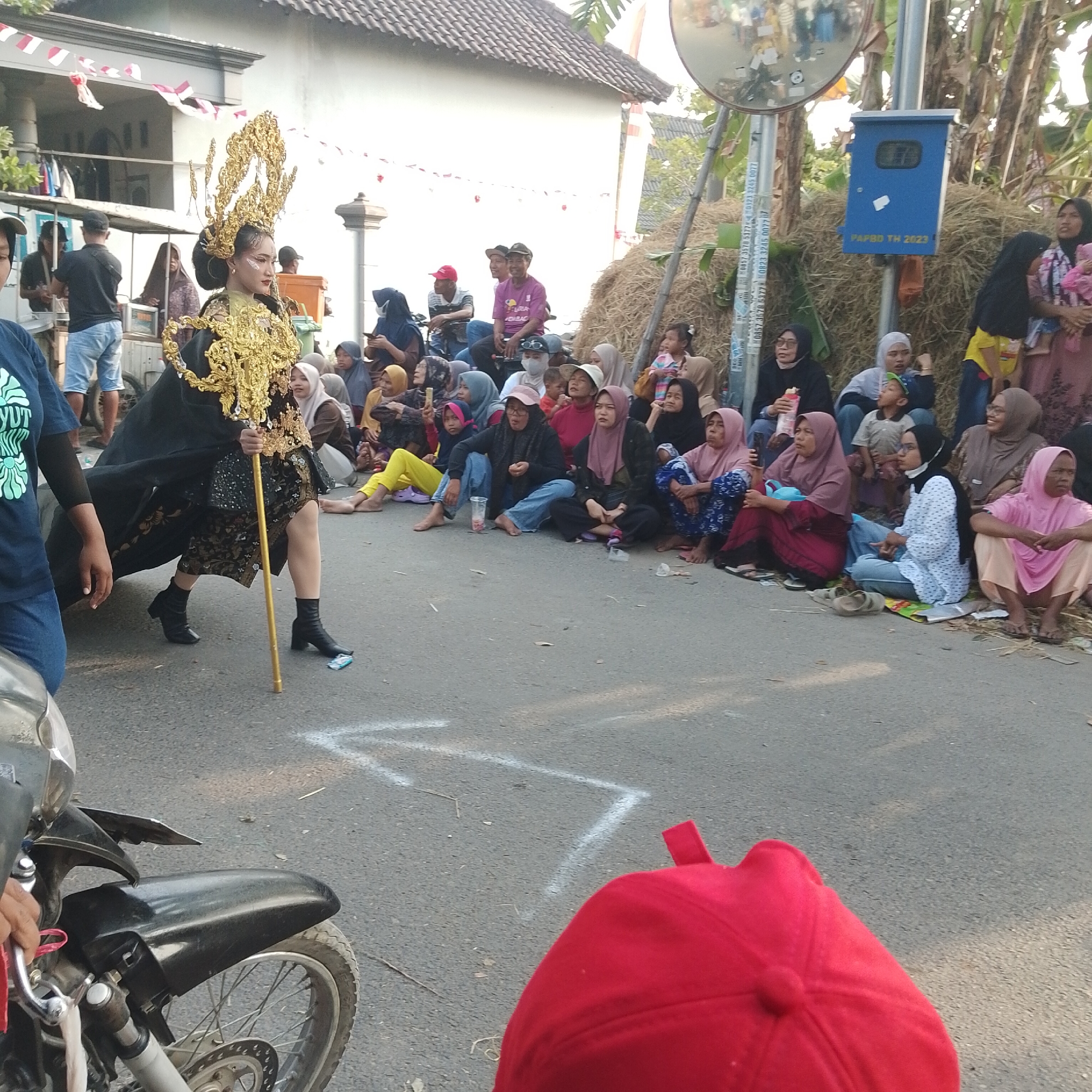 The image depicts a street scene in Palang, Indonesia, likely during a local festival or parade. In the foreground, a young woman dressed in a traditional ornate costume, complete with a golden headdress and a flowing black cape, is walking while holding a golden staff. She is wearing black ankle boots. Her face is turned to the side, with makeup applied.
Surrounding the central figure are numerous spectators seated or standing on the side of the road. Many of the spectators are women and children, most of whom are wearing headscarves or hijabs in various colors. Some are holding cell phones, suggesting they are capturing the event. The background includes buildings with tiled roofs decorated with red and white banners, a pile of hay bales, a blue utility box labeled "PAPBD TH 2023," and trees. A motorcycle is partially visible in the lower left corner of the image, with a red object, possibly a cap or bag, in the bottom right corner. White markings are painted on the asphalt road, indicating a designated path or area. The overall atmosphere appears to be festive and communal.