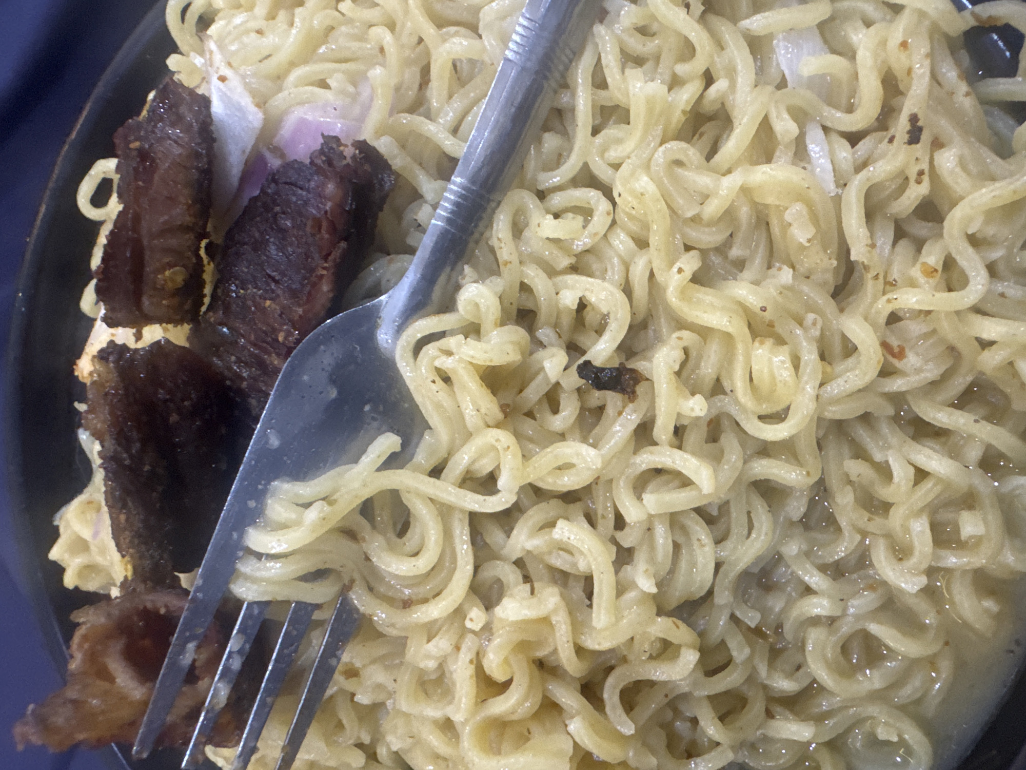A close-up overhead shot shows a serving of instant noodles in a black bowl. The noodles are a light yellow color and appear to be coated in a sauce. Several pieces of dark brown, grilled meat are scattered on top of the noodles, along with some white and purple onion slices. A metal fork is placed diagonally across the noodles, with its tines embedded in the dish. The background is a dark blue surface, possibly a tablecloth or a table. There is no indication of the city of Abuja, Nigeria in the image.