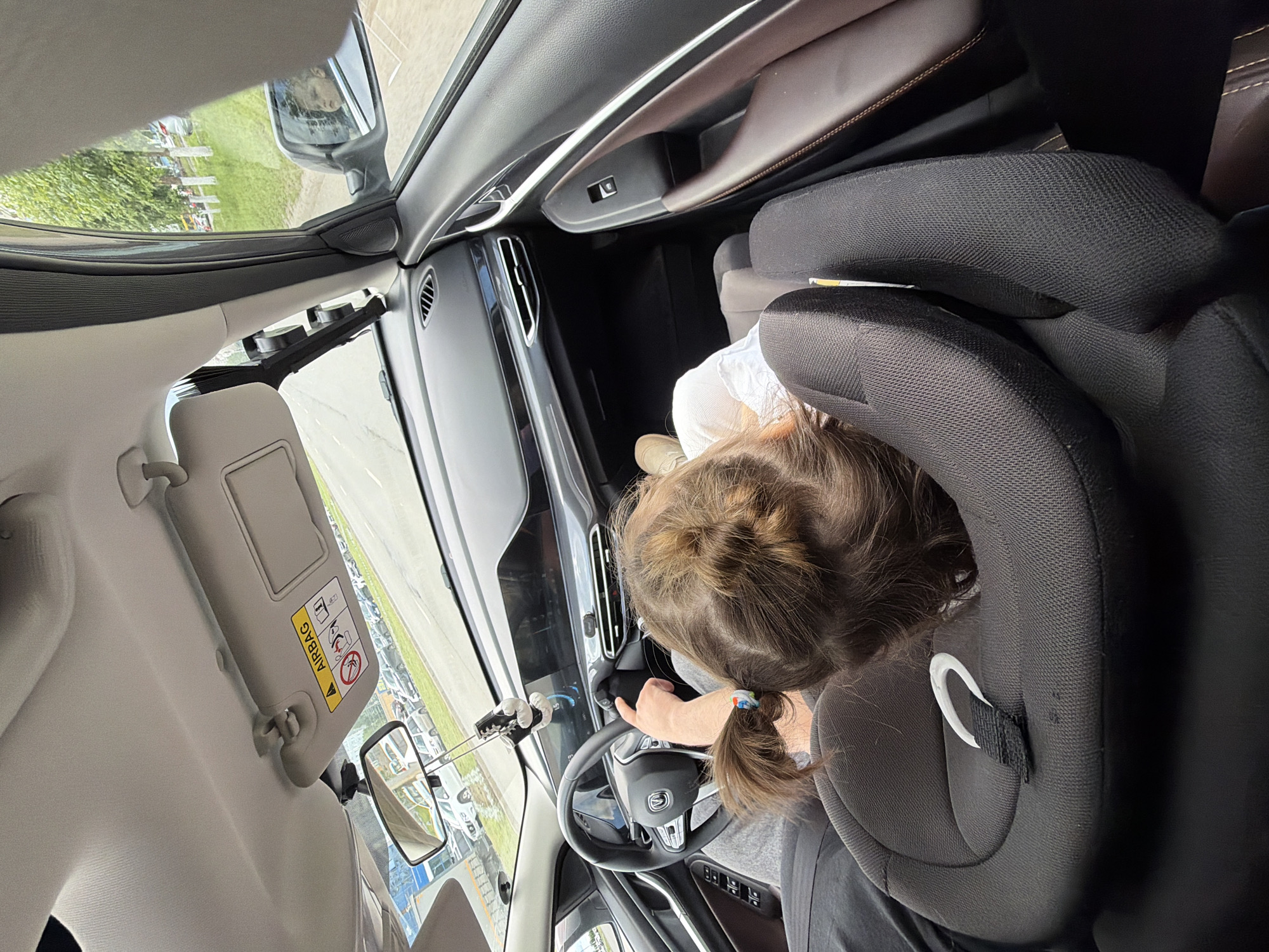 The image shows the interior of a car, looking forward from the back seat where a young child is seated in a car seat. The child is facing away from the camera, with their back to the viewer, and has brown hair tied in a ponytail. They are wearing a white shirt and grey pants. In the foreground, the black car seat is prominent. The dashboard of the car is visible, featuring a large digital display and a steering wheel. The driver's hand is on the steering wheel. Through the windshield and side windows, a road with other vehicles and trees is visible, suggesting the car is in motion. A sun visor with an "AIRBAG" warning sticker is visible overhead. The interior of the car appears modern, with brown leather accents on the doors and dark grey or black upholstery. The scene is likely captured in Izhevsk, Russia, given the provided context.