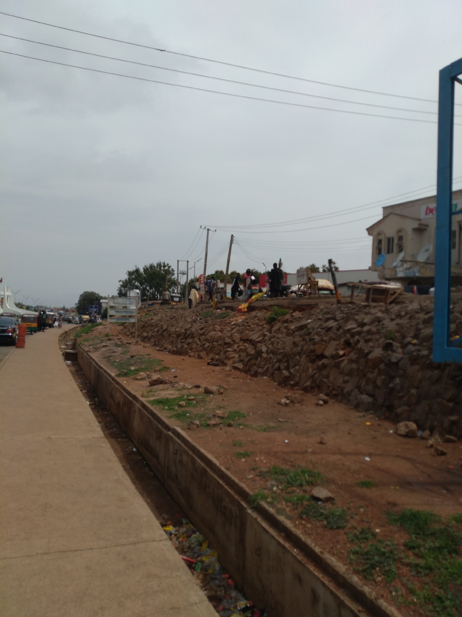The image depicts a street scene in Jos, Nigeria, under an overcast sky. A concrete sidewalk runs along the left side of the frame, bordering a shallow, concrete-lined drainage ditch filled with debris and trash. Beyond the ditch, a rocky embankment rises, topped with a scattering of dry grass and some larger rocks. Further back, a dirt area shows signs of being a marketplace or gathering space, with several wooden structures and makeshift stalls visible. A few people are present in this area, some standing and some possibly interacting or engaged in commercial activities. In the background, buildings line the street, including a light-colored, multi-story structure with windows and a sign that appears to say "bet" in red. Utility poles and overhead wires crisscross the sky. To the far left, vehicles are parked along the street, suggesting ongoing traffic or activity. The overall impression is one of an urban environment with visible signs of development and economic activity, but also challenges with sanitation and infrastructure.