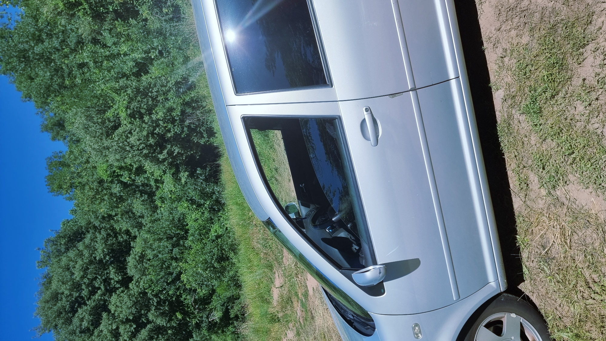 The image shows a silver four-door car parked on a dirt and grass area. The car's right side is visible, with the front and rear passenger doors and windows in focus. The windows are tinted, reflecting the surrounding trees and blue sky. A line of dense green trees forms the background. The car appears to be in a rural or natural setting, possibly near Montagut i Oix, Spain, given the lush vegetation. There are no people visible in the image. The car is angled, suggesting it is parked on a slight incline or on the side of a road. The overall scene is bright and sunny, indicating daytime.