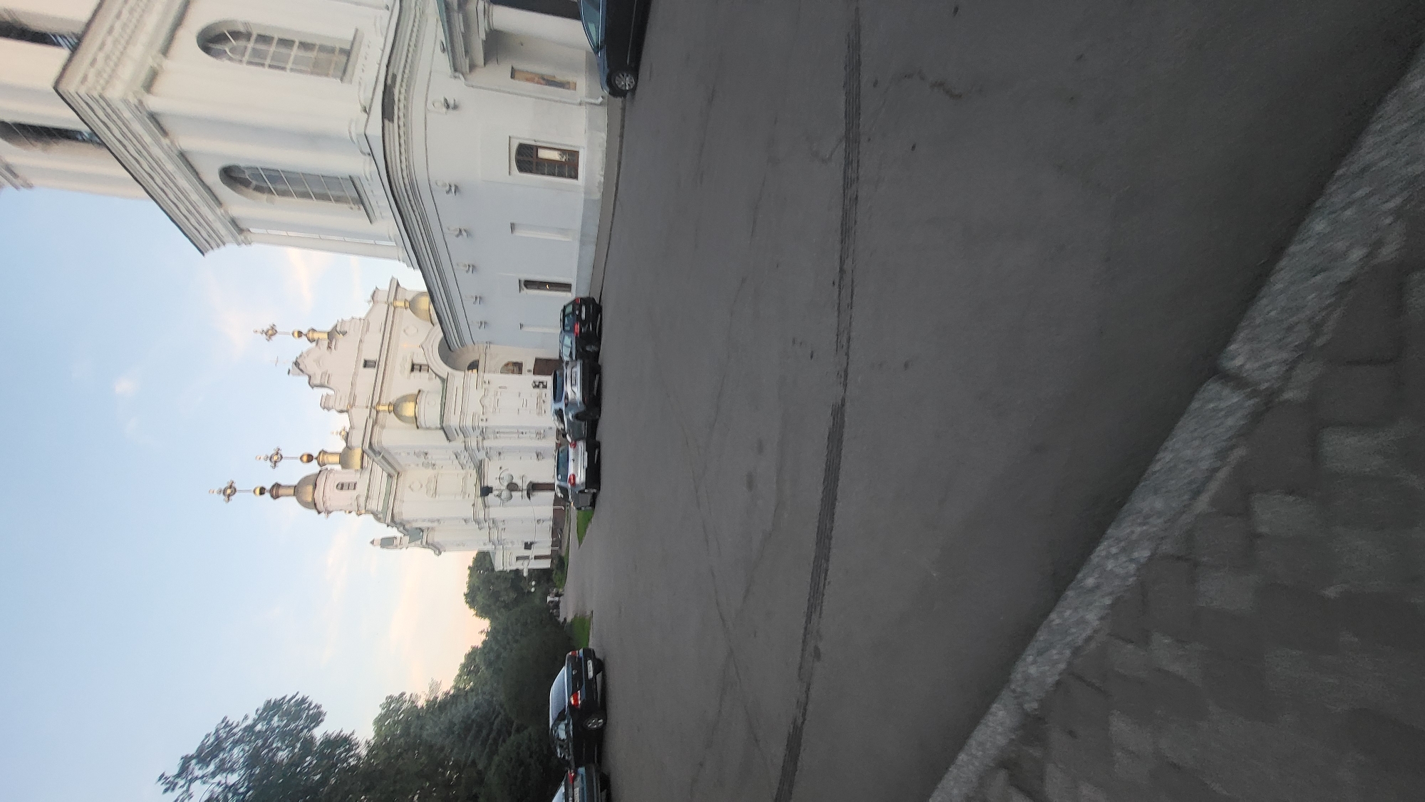 An exterior, eye-level, medium shot captures a paved area in front of a large, ornate white building with gold-domed towers, likely a church or cathedral, under a pale sky. Several cars are parked on the asphalt, including dark sedans and SUVs. To the left of the frame, a dense line of dark green trees forms a natural border. The lighting suggests it is late afternoon or early evening.