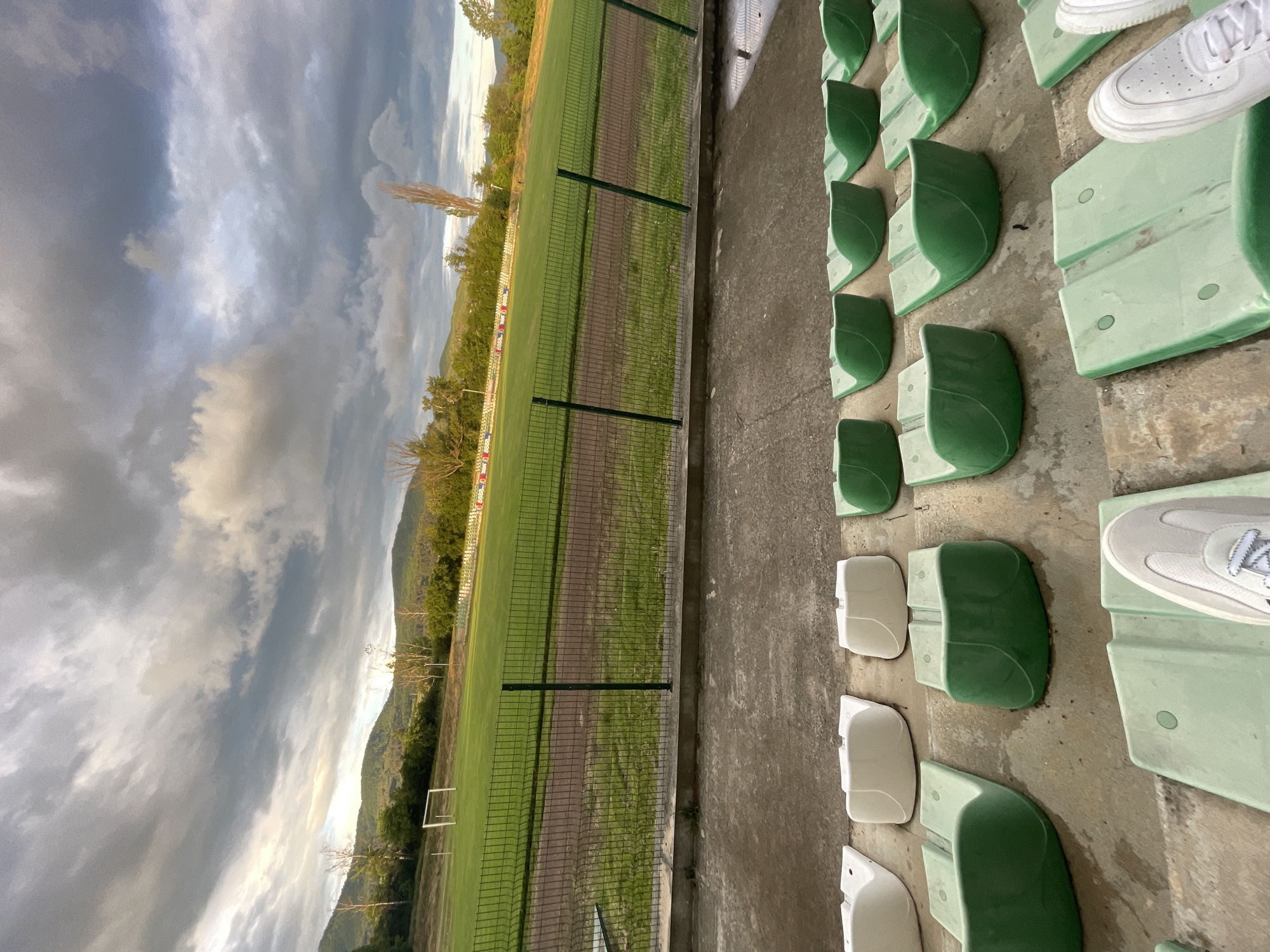 The image presents a view from the stands of a sports field, likely a soccer pitch, under a cloudy sky. In the immediate foreground to the right, rows of stadium seats are visible, predominantly in a light green color, with some white seats interspersed. These seats are fixed onto a concrete floor. On two of the green seats, a pair of white athletic shoes (specifically, white Nike Air Force 1s) and a pair of light grey athletic shoes are resting, indicating a recent or imminent human presence, though no people are depicted.
A green wire mesh fence separates the spectator area from the playing field. The field itself is covered in green grass, with some areas appearing more worn or showing patches of darker earth. A white soccer goal is visible on the far side of the field. In the background, rolling mountains covered with dense greenery frame the setting.
The sky is largely overcast, displaying various shades of grey clouds, with some brighter patches suggesting that it is daytime, possibly late afternoon or early morning. No specific activity or event is taking place on the field, which appears empty. Along the far edge of the field, beyond the goal, there are advertising hoardings with indistinct, repeated red and white patterns or logos, possibly including numbers like '8888', but the text is not clearly legible. The scene conveys a quiet, contemplative atmosphere. The location context provided is Ihtiman, Bulgaria.