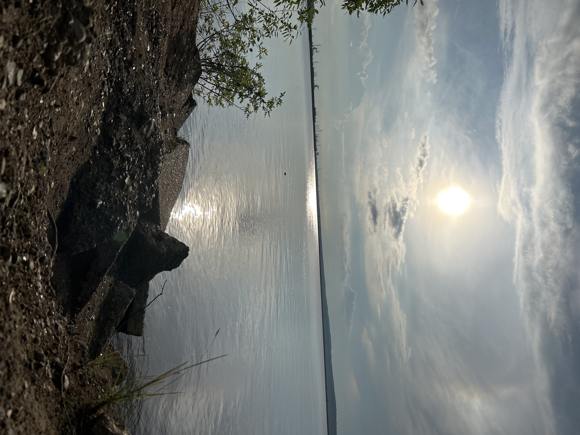 A horizontal, high-angle, low-light photograph shows a serene body of water, likely a reservoir or lake, on a clear day. The sun, a bright orb, is positioned in the upper right quadrant of the image, casting a shimmering reflection on the water's surface. Wispy clouds stretch across the sky, adding depth and texture to the scene. A small, dark object, possibly a buouy or a duck, can be seen in the middle of the water.
In the foreground, the rough texture of a rocky embankment is visible, with sparse vegetation clinging to its surface. The embankment appears to be man-made, possibly a reinforced bank or a section of a dam. The overall atmosphere of the image is peaceful and tranquil, evoking a sense of natural beauty and solitude. The time of day appears to be late afternoon or early evening, judging by the sun's position and the soft lighting. The location is identified as Chaikovsky, Russia, which is known for its large hydroelectric dam and reservoir on the Kama River.