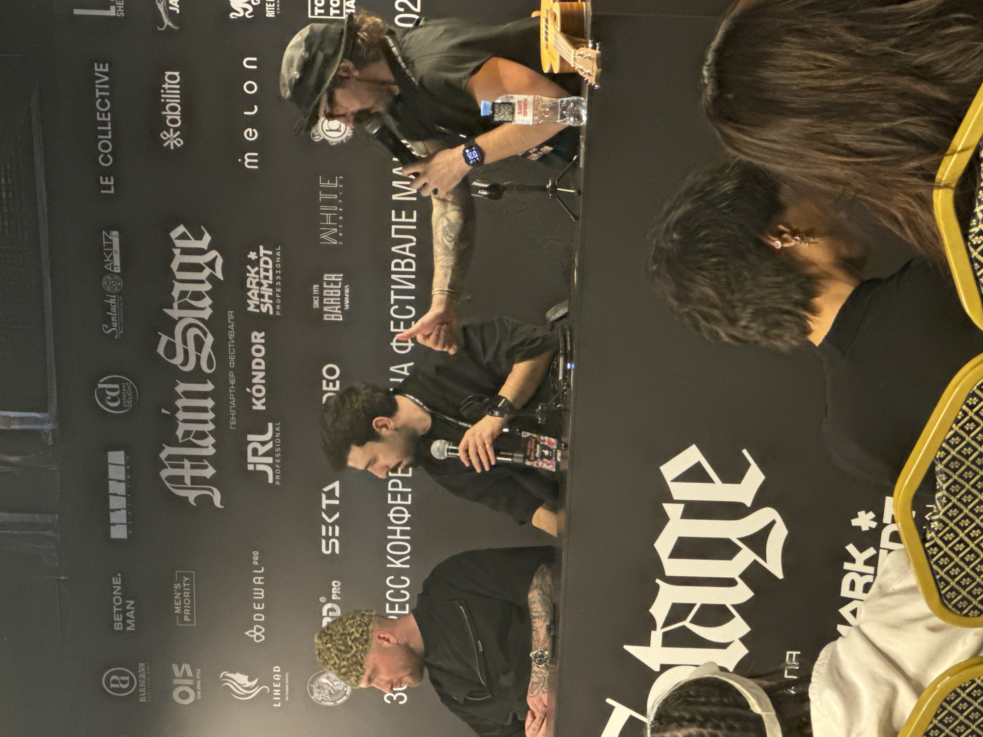 The image shows a panel of three individuals sitting behind a table at an event. The background features branding for various companies, including "Main Stage," "JRL Kondor," "Barber Marmara," and others. The panelists appear to be engaged in a discussion, with microphones positioned in front of them. A water bottle is visible on the table. In the foreground, parts of the audience can be seen, adding context to the setting.
