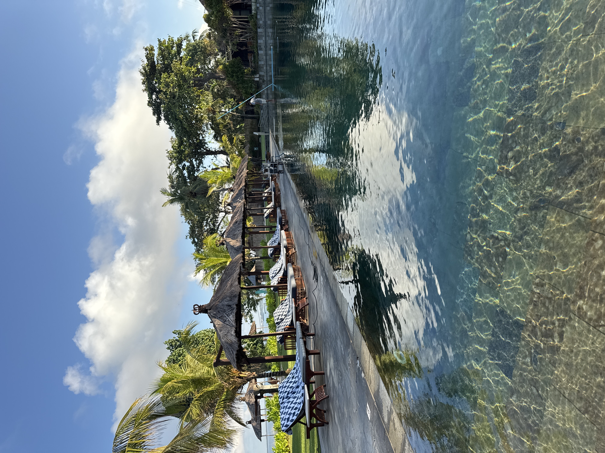 The photograph depicts a tranquil scene at a resort in Jimbaran, Indonesia. Along the edge of a clear, blue-water pool, several lounge chairs are arranged under thatched umbrellas. The chairs are partially covered with patterned fabric. Lush tropical foliage and trees line the edge of the pool area. The sky is blue with scattered, fluffy white clouds, which are reflected in the pool's surface. The pool's shallow end appears to be tiled, with sunlight creating shimmering patterns on the pool floor. No people are visible in the immediate scene.