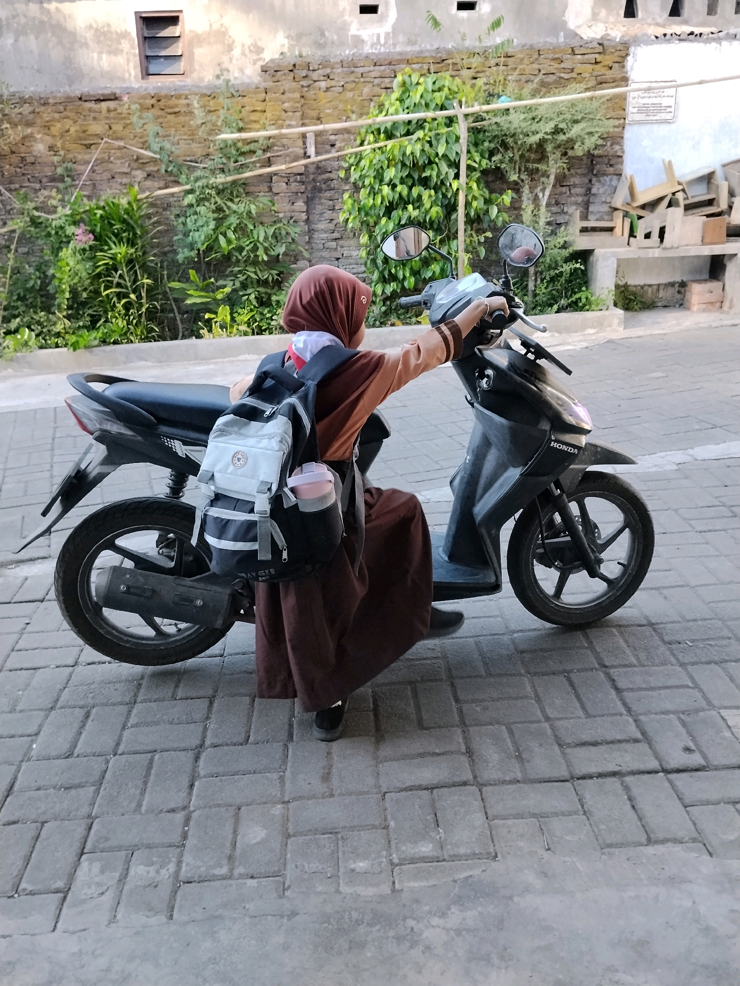 A person wearing a brown hijab and a brown dress is sitting on a black Honda scooter in a city street in Kejawan, Indonesia. The person has a backpack on their back with a water bottle attached to the side. The street is paved with brick. In the background, there is a stone wall and some greenery. A window with louvers is visible on the wall. There are also some wooden crates and chairs stacked in the background. The scene appears to be outdoors in daylight.