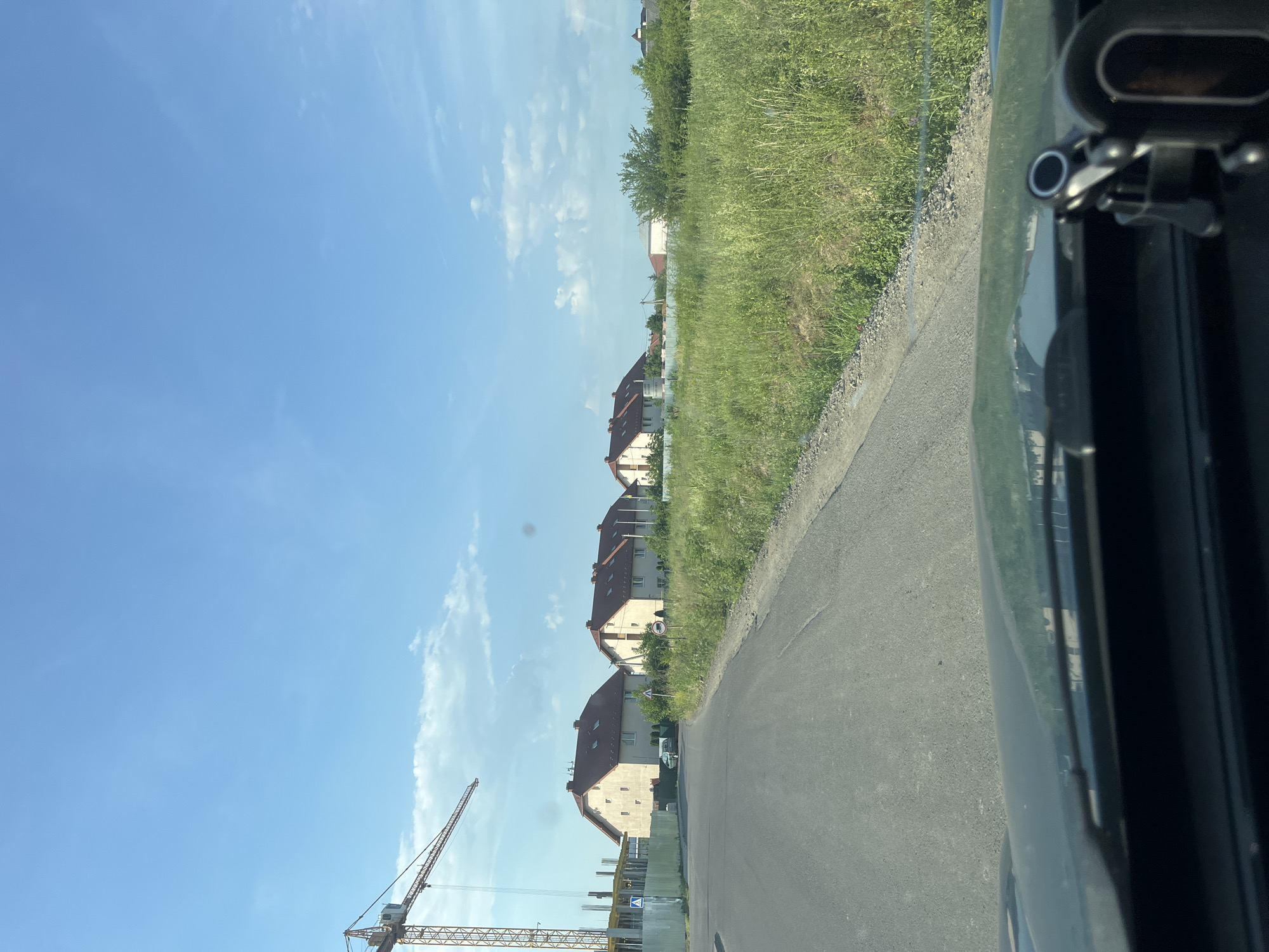 This is a photograph taken from inside a vehicle, looking out at a rural or suburban landscape in Mynai, Ukraine. The scene is dominated by a clear blue sky with scattered white clouds. In the mid-ground, a construction crane is visible on the left side of the image, suggesting ongoing development. Adjacent to the construction site, a row of identical, light-colored houses with red tiled roofs are situated. These houses appear to be relatively new and are spaced apart. A paved road curves through the foreground, leading towards the houses. To the right of the road is a dense area of tall, green grass and vegetation. The overall impression is one of a developing residential area on the outskirts of a town or city, under a bright, sunny sky.