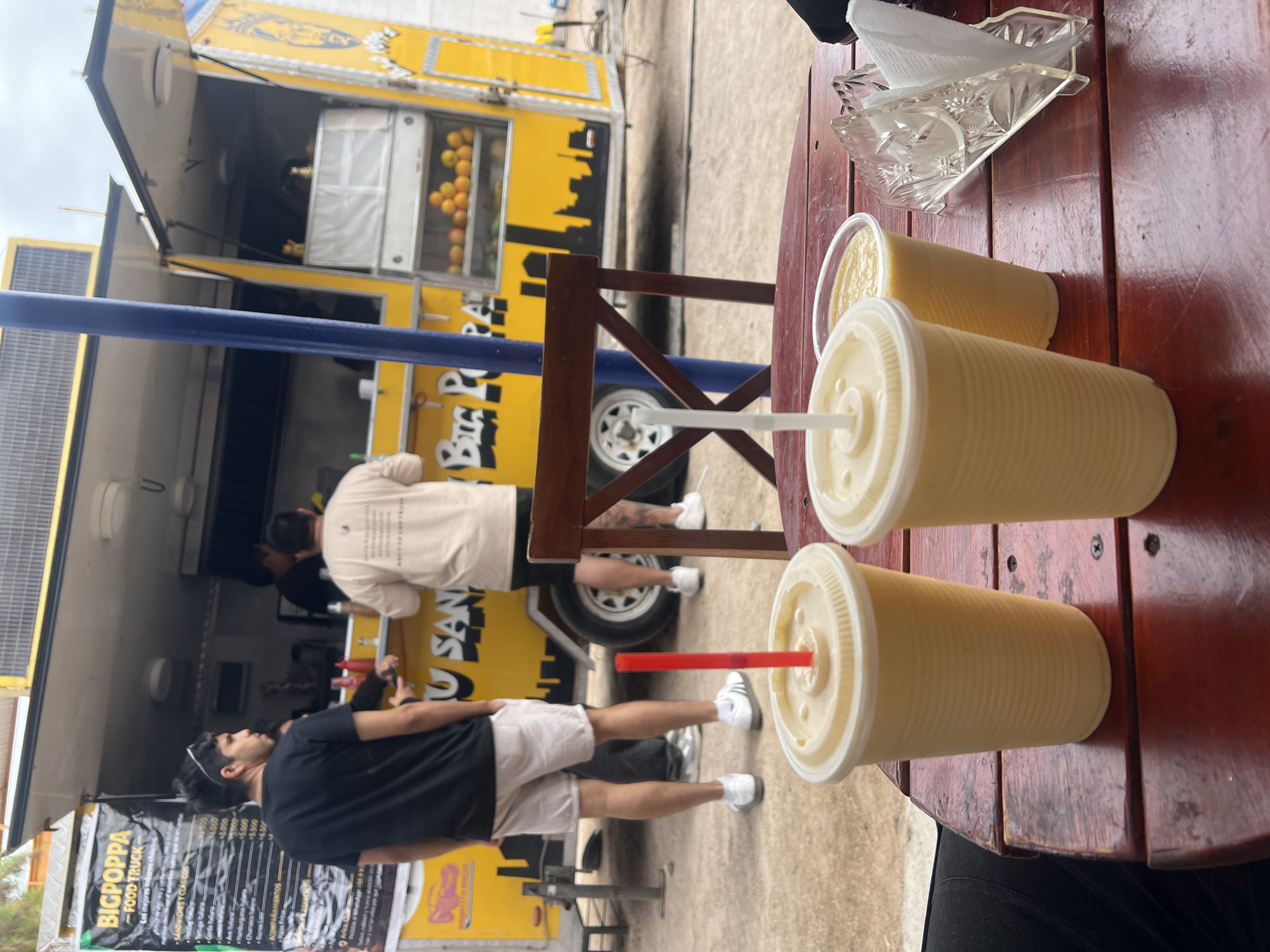 The image shows an outdoor dining scene, likely in Arica, Chile. In the foreground, three large, opaque yellow drinks with lids and straws sit on a wooden table alongside a clear plastic napkin holder filled with white napkins. In the background, partially obscured by the blue support pillar of a food truck, are two men standing in front of the yellow food truck, "BIGPOPPA." The truck's signage, in both English and Spanish, lists sandwiches and "chilenos" food, with prices displayed in Chilean pesos. One man wears a white shirt with writing on the back and dark shorts, while the other wears a black shirt and light shorts. The truck is decorated with a colorful image and text. The area surrounding the table and truck appears to be a paved outdoor space.