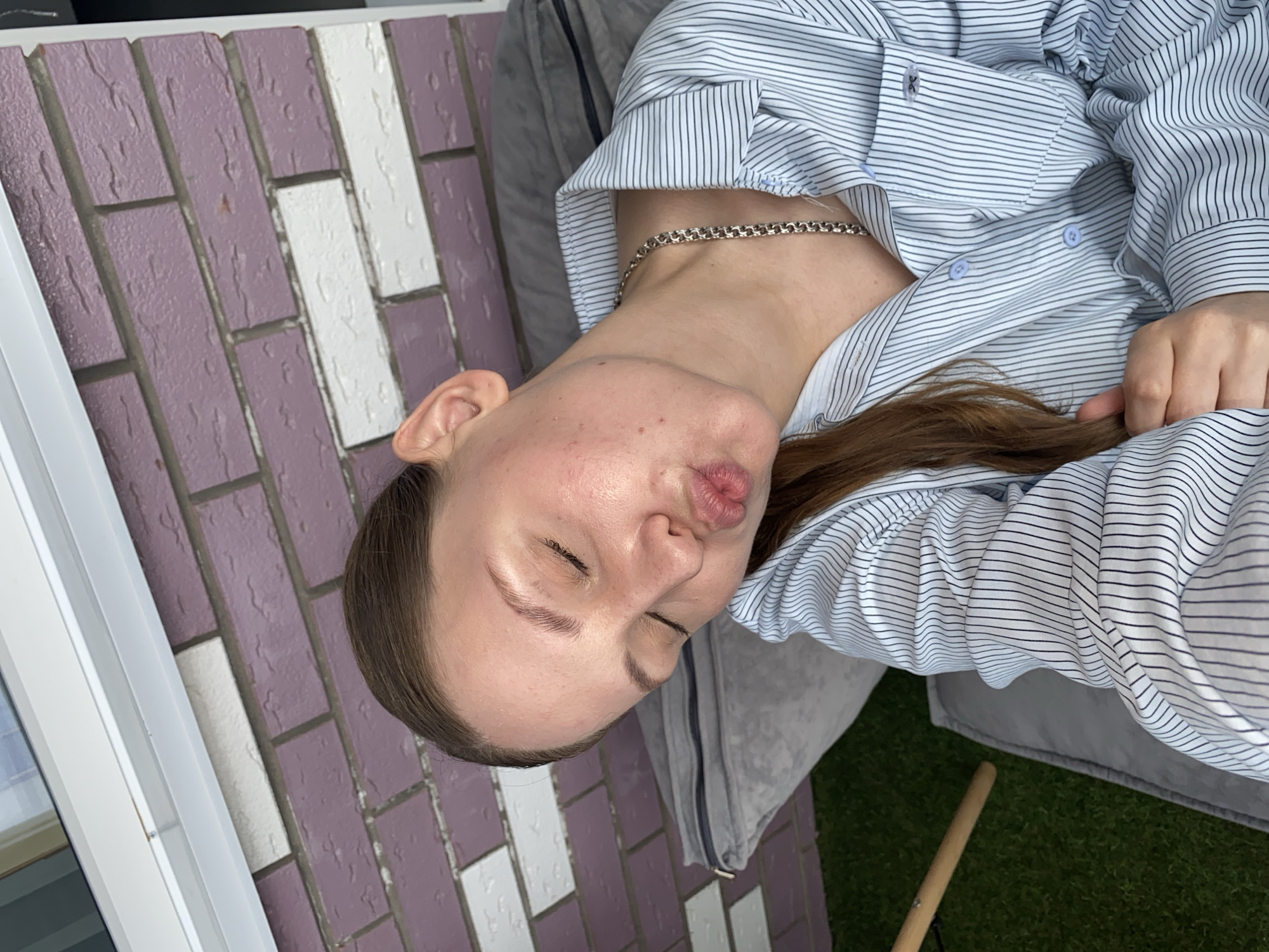 A young Caucasian woman with brown hair is pictured from the chest up, taken outdoors in what appears to be a city setting, possibly Izhevsk, Russia. She is wearing a light blue and white striped button-down shirt and a silver chain necklace. Her eyes are closed, and her lips are pursed in a kissing gesture, indicating a selfie. She is leaning back against a grey cushion. In the background, there is a purple and white brick wall and a glimpse of artificial green turf and a wooden object. The lighting suggests it is daytime.