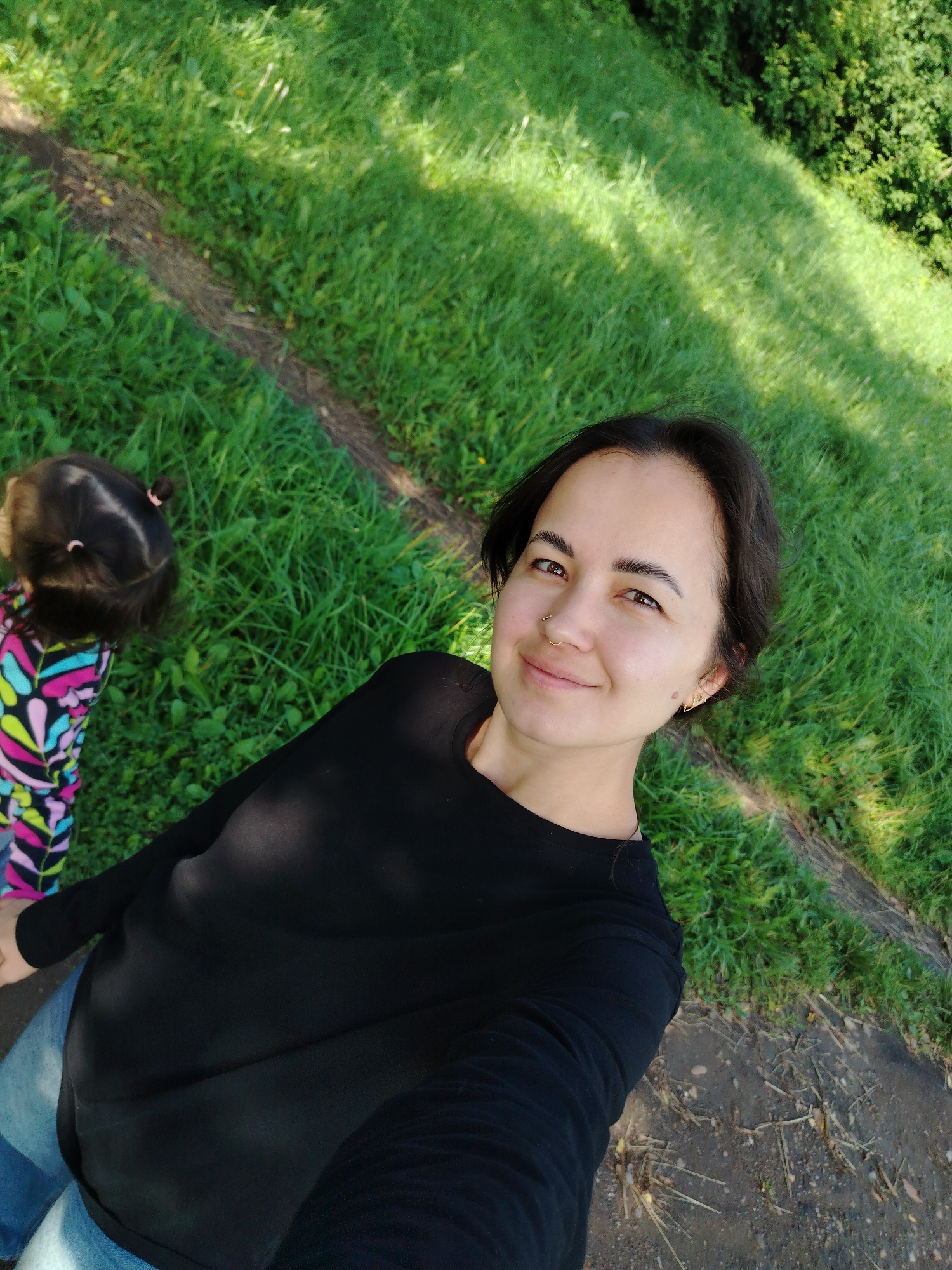 A medium shot captures a woman in a black long-sleeved shirt and blue jeans posing for a selfie outdoors, with a young child in a colorful patterned outfit visible to her left. The woman has short dark hair, brown eyes, and is wearing a nose ring and earrings. She is smiling and looking at the camera. The child, with dark hair styled in two pigtails, is facing away from the camera and looking towards the left. They are both situated on a grassy slope, with a path running horizontally behind them. The environment appears to be a park or a green space within the city of Kuyeda, Russia. The lighting suggests it is a sunny day.