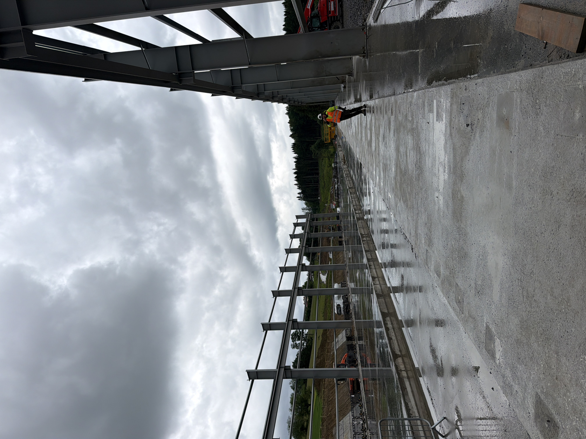 This image depicts a construction site on a cloudy day. In the foreground, a wet concrete walkway runs alongside a large, unfinished steel structure. A person wearing a high-visibility vest and hard hat is standing on the walkway, appearing to be talking on a phone. In the mid-ground, there are more steel beams, safety barriers, construction equipment including an excavator and a truck, and a sloped area with some vegetation. The background shows a line of trees and a hilly landscape. The presence of puddles and reflections on the walkway indicates recent rainfall. The scene is consistent with a construction project in an outdoor, potentially rural, environment.