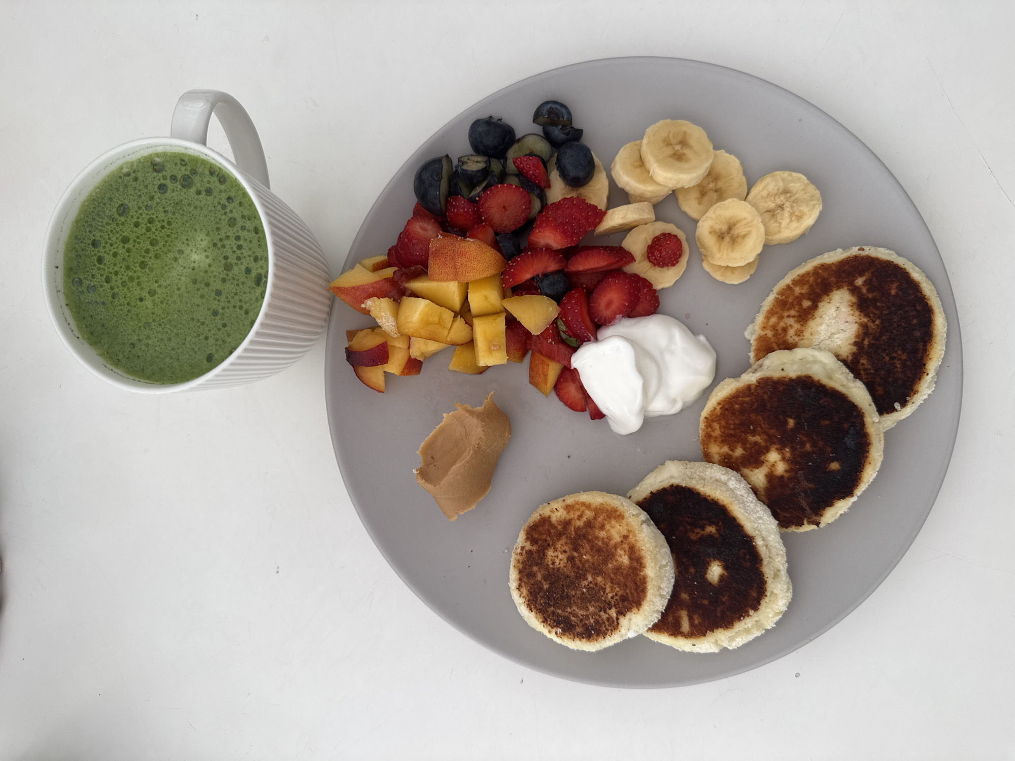 A top-down shot shows a light gray plate filled with food placed on a white surface, likely a table. The plate contains a portion of cubed peaches, a mix of blueberries and sliced strawberries, sliced bananas (some with strawberry pieces on top), a dollop of white yogurt, a smear of peanut butter, and four small, round, browned pancakes or cottage cheese pancakes (syrniki). A mug filled with a green, frothy liquid (possibly matcha or another green beverage) sits to the left of the plate. The scene is simple and appears to represent a breakfast or brunch meal. No further location information or actions are present.