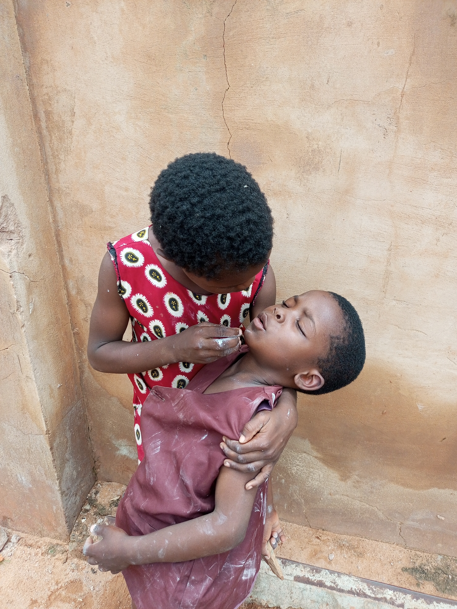 Two young children are positioned in front of a textured, beige-colored wall with visible cracks. The child on the left, wearing a red and white patterned dress, is leaning forward and appears to be administering something, possibly medicine, to the child on the right. The child on the right, wearing a dark reddish-brown dress, is lying on their side, eyes closed, with their head supported by the other child's arm. Their bodies are covered in a fine white powder. The ground beneath them is dirt with some scattered debris. The overall scene suggests a moment of care or gentle interaction between the two children. The location is specified as Tempure, Nigeria.