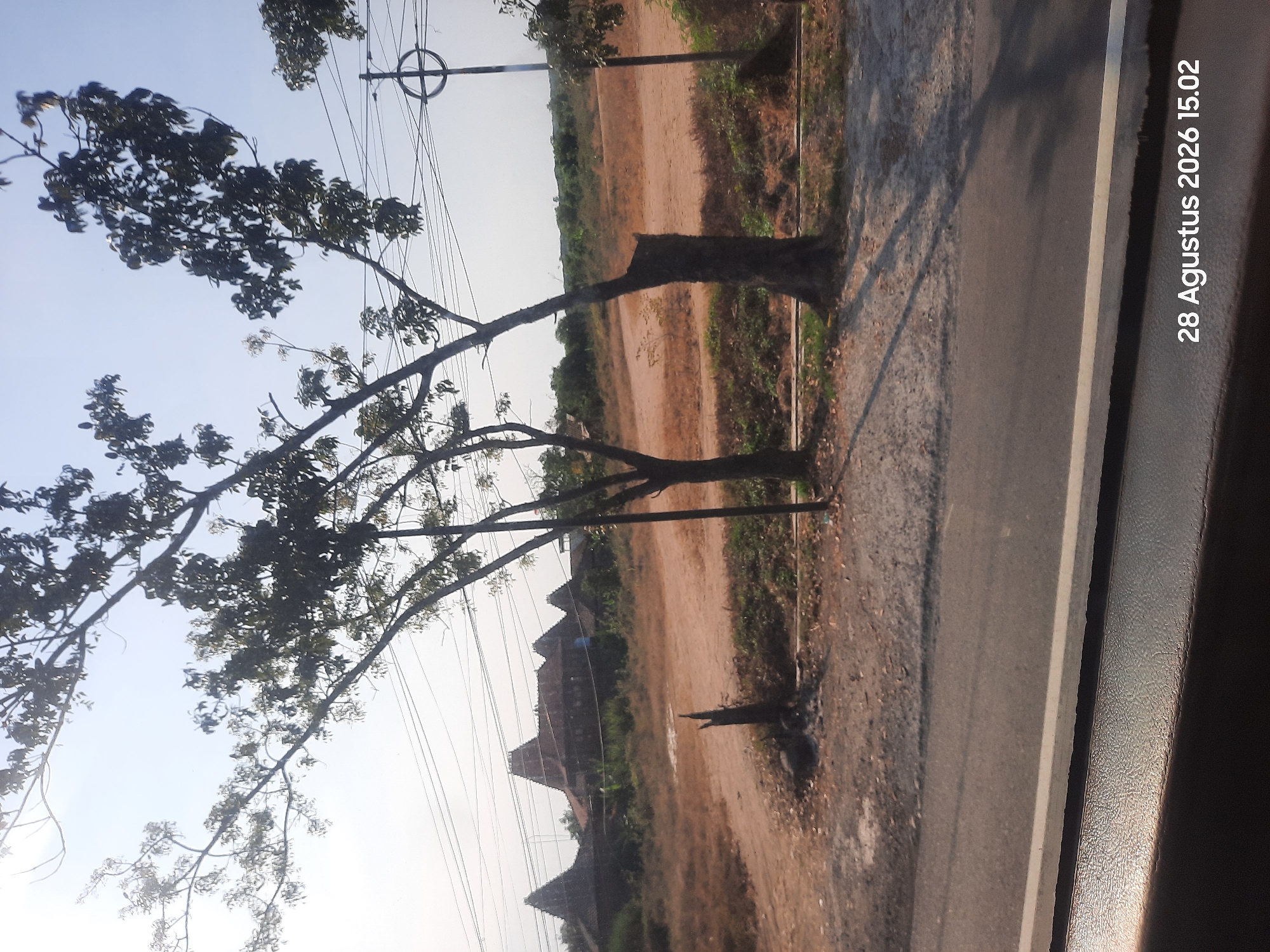 The image shows a rural landscape with a dirt road in the foreground and a line of traditional Indonesian houses with distinctive pitched roofs in the background. There are several trees with sparse foliage, casting long shadows on the ground, suggesting it is late afternoon. Power lines are visible strung between poles, indicating a connection to the grid. The sky is a pale blue with some hazy clouds. The date stamp on the image reads "28 Augustus 2026 15.02", implying the photograph was taken in Bulang, Indonesia, at 3:02 PM on August 28, 2026. The scene appears to be undisturbed and quiet, with no people or animals visible.